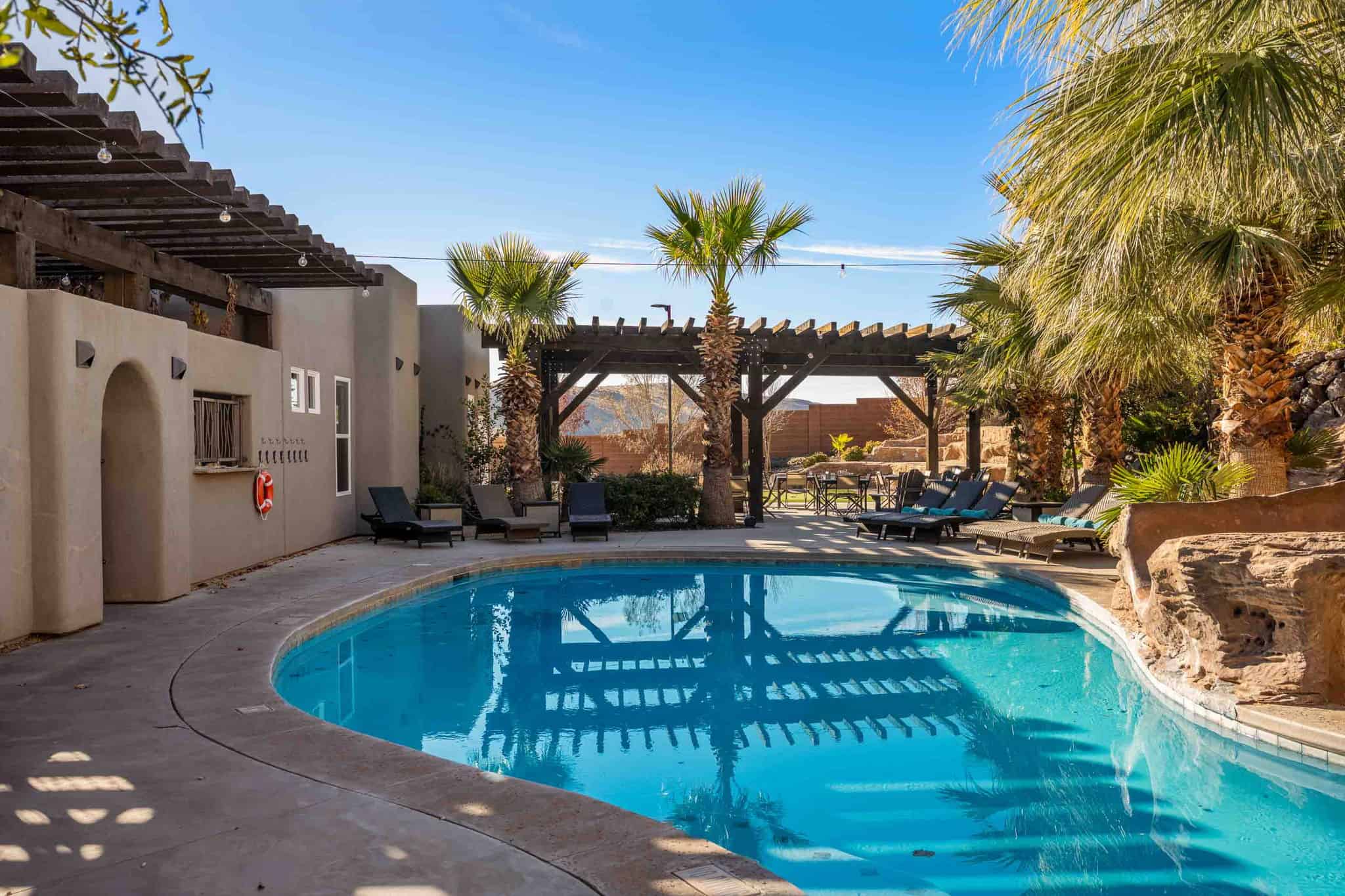 Curved swimming pool with integrated rock formation, surrounded by established palm trees, an outdoor bar area, and multiple wooden pergolas
