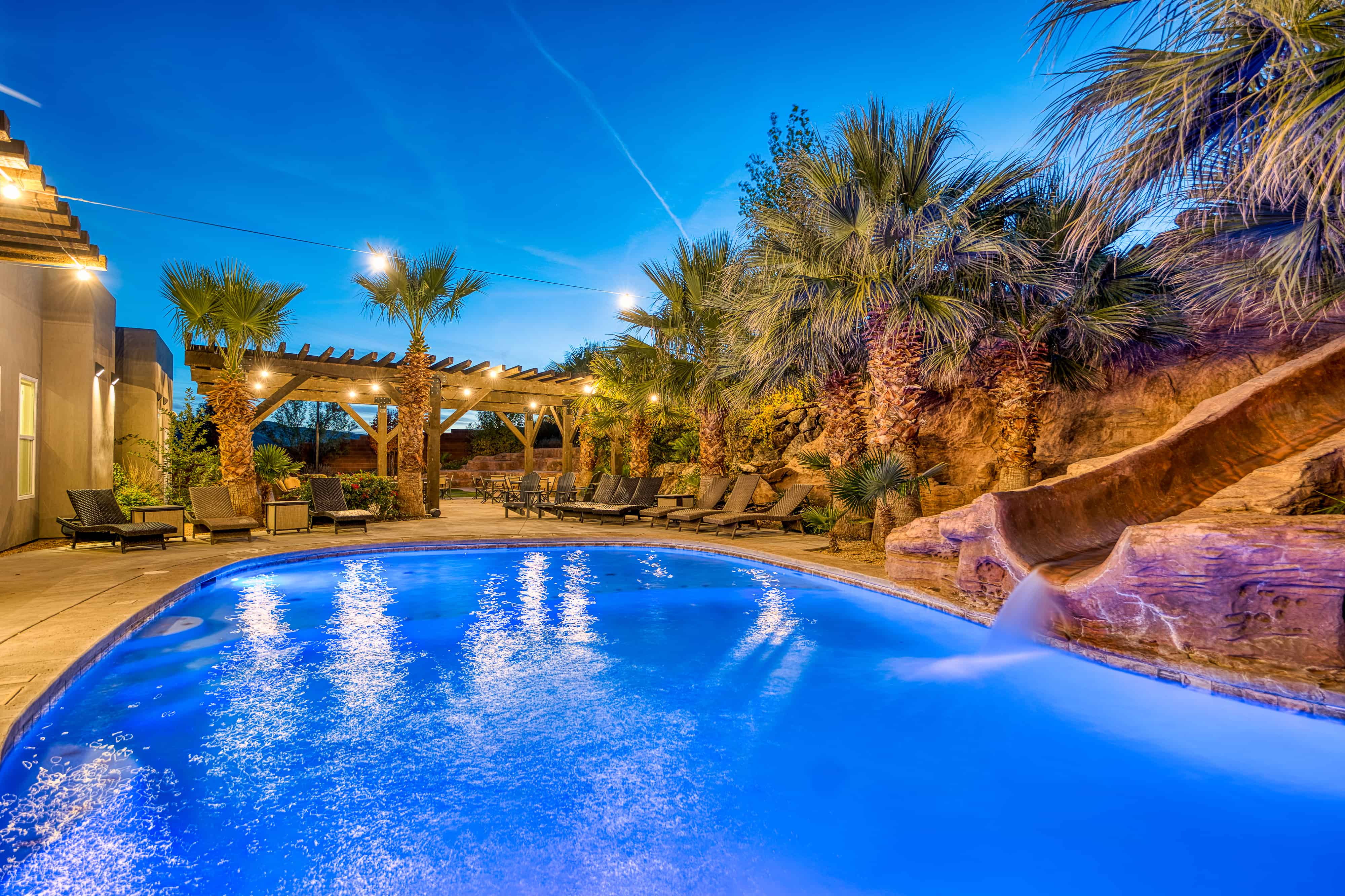 Resort-style backyard featuring a large swimming pool, rock water slide, pergola-covered patio, and mature palm trees