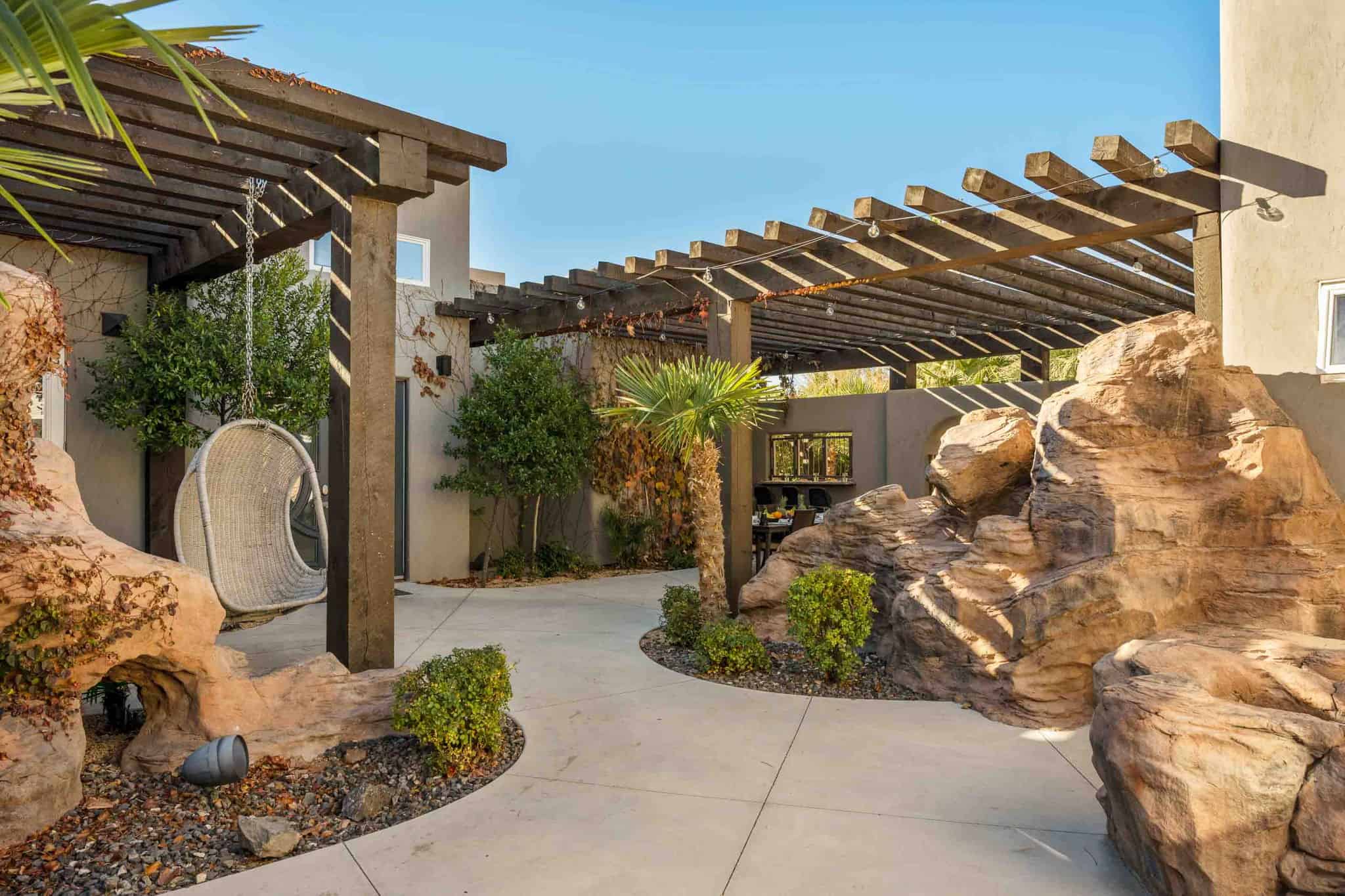 Outdoor living area featuring a large wooden pergola, concrete hardscaping, artificial rock formations, mature palm trees, and integrated landscaping