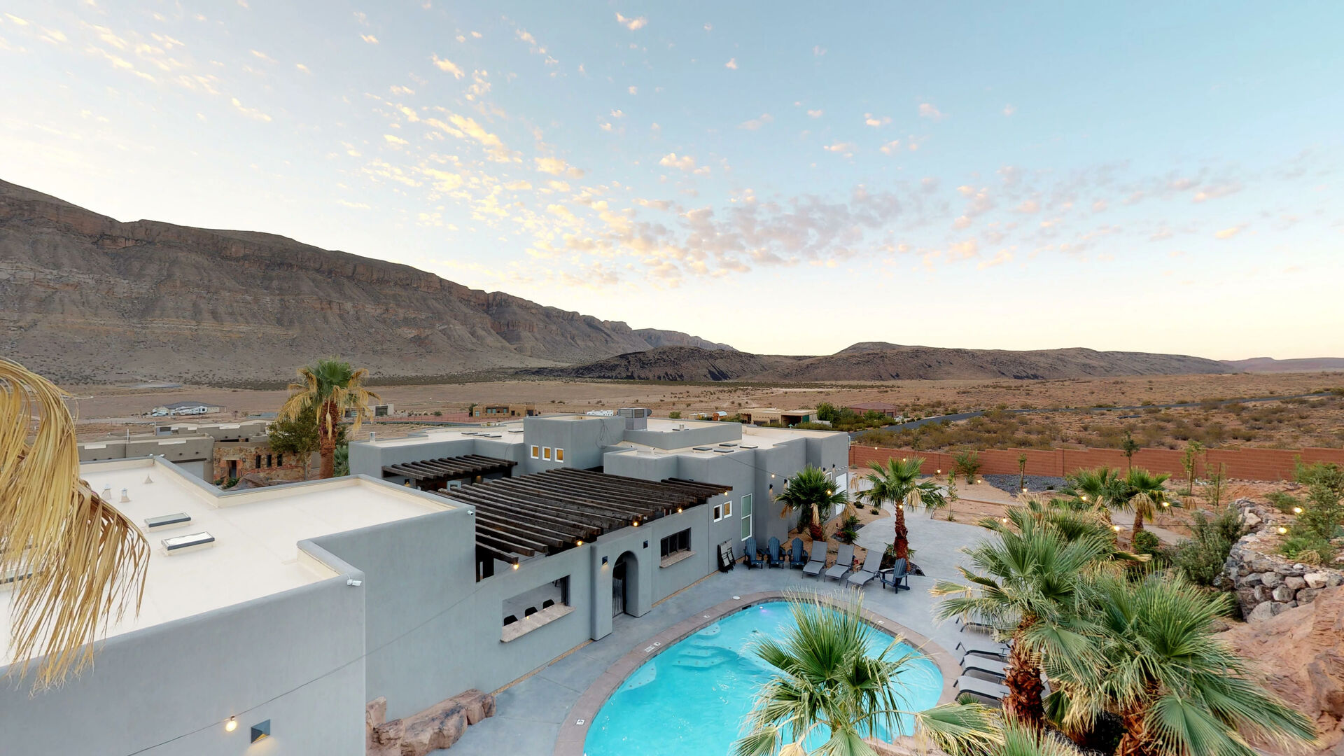 Desert landscape featuring a modern stucco exterior, a large swimming pool, a pergola-covered patio, and mature palm trees