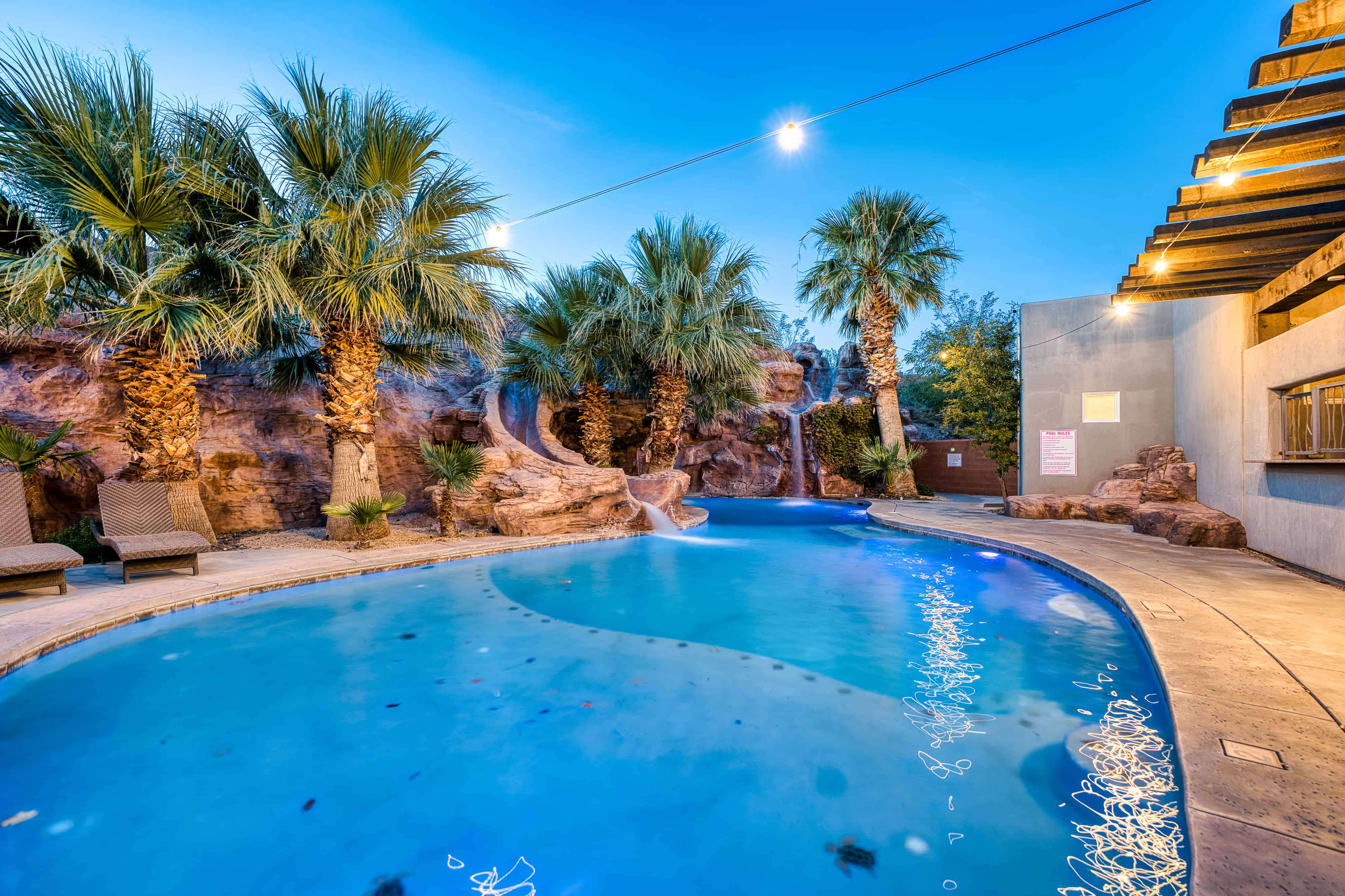 Curved swimming pool featuring a rock waterfall, surrounded by mature palm trees and concrete decking