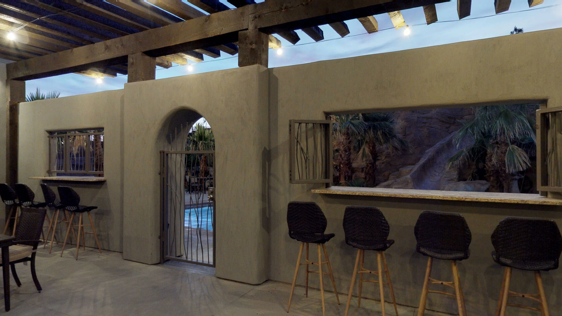 Exterior patio featuring an open pergola with string lighting, stucco walls, a service window with a counter, and an arched entryway with a wrought iron gate leading to a pool area