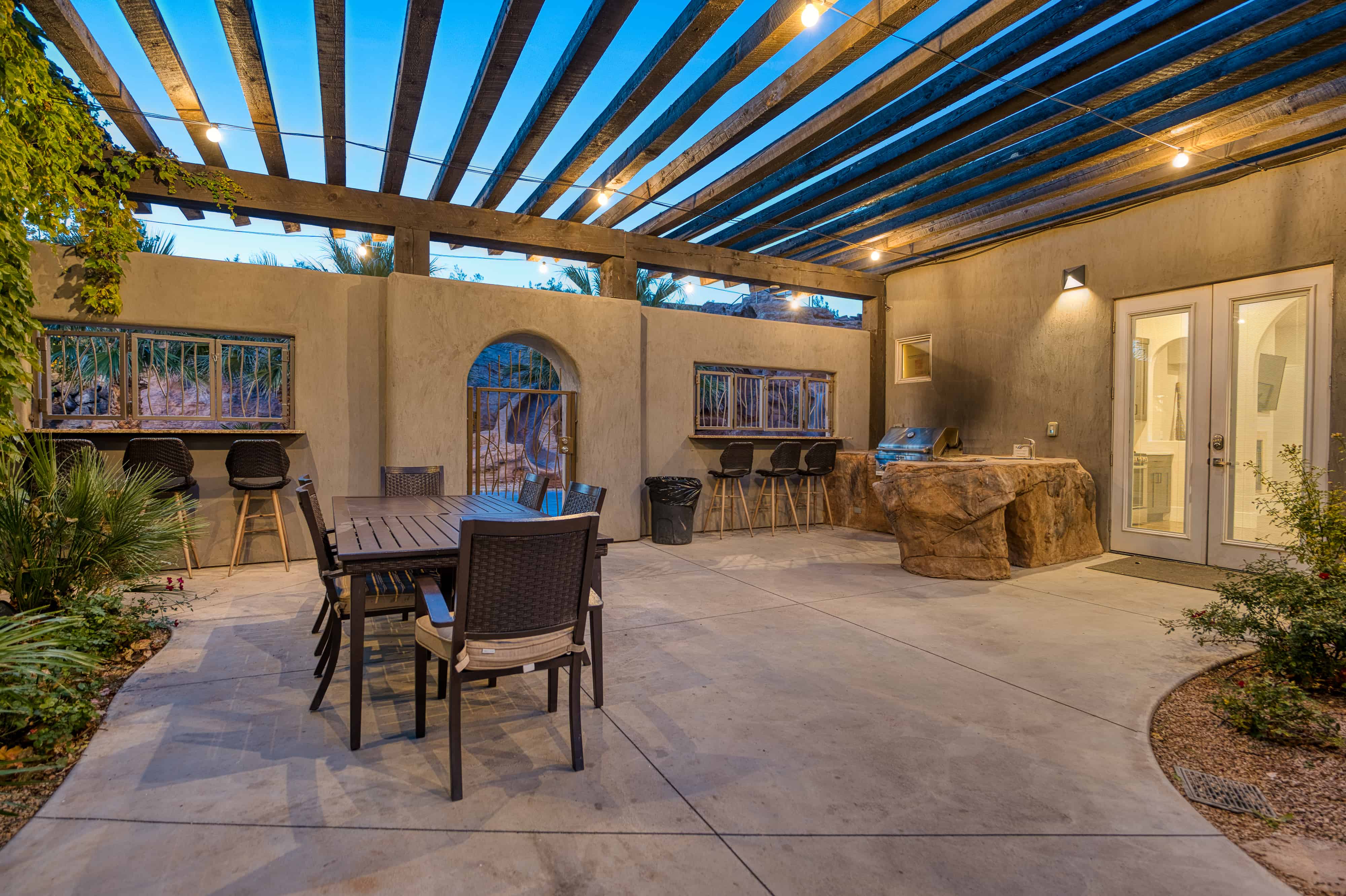 Expansive concrete patio featuring a wood pergola with string lighting, a built-in outdoor bar area, and a rock-finish outdoor kitchen station