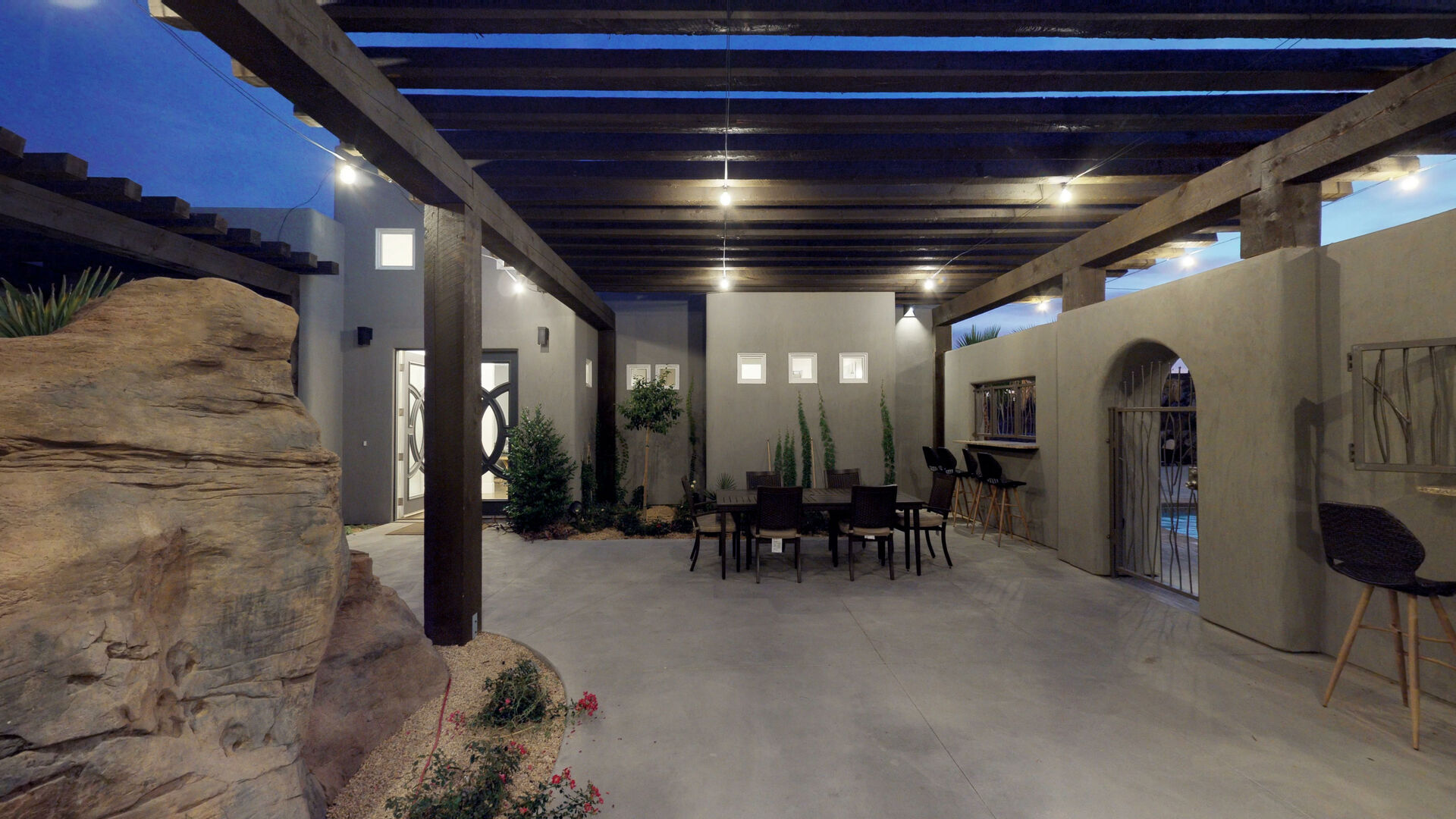 Expansive outdoor patio featuring a wood beam pergola with integrated lighting, concrete flooring, and a rock feature