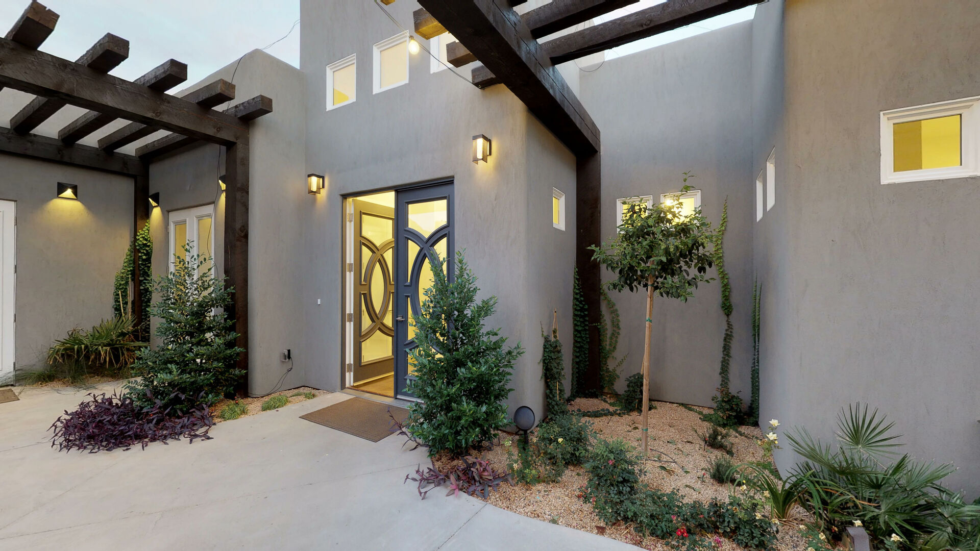 Contemporary stucco exterior featuring an ornate glass-paneled entry door, dark wood pergolas, exterior wall sconces, and low-maintenance landscaping with mature trees