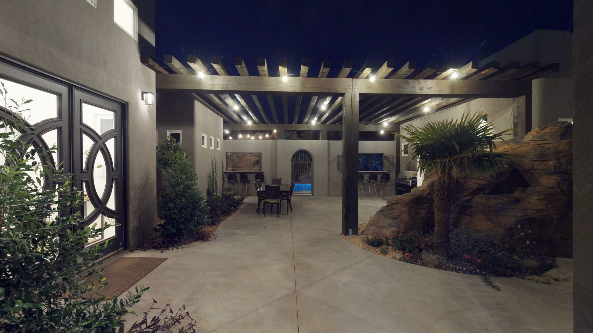 Expansive outdoor patio featuring a wood pergola with string lighting, a stucco exterior, and a decorative double entry door with circular ironwork