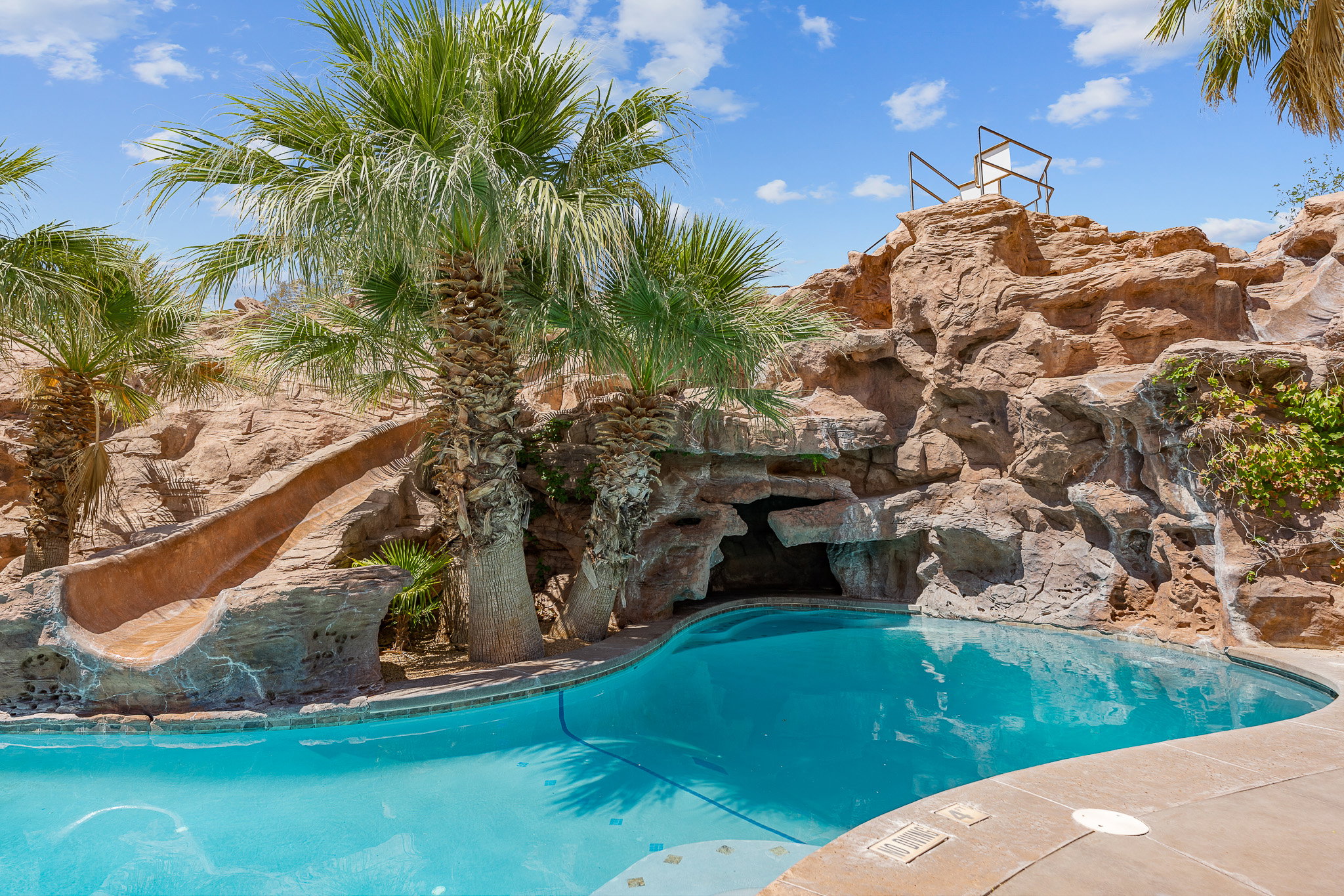Custom-shaped swimming pool featuring a rock grotto, integrated water slide, concrete decking, and mature palm trees