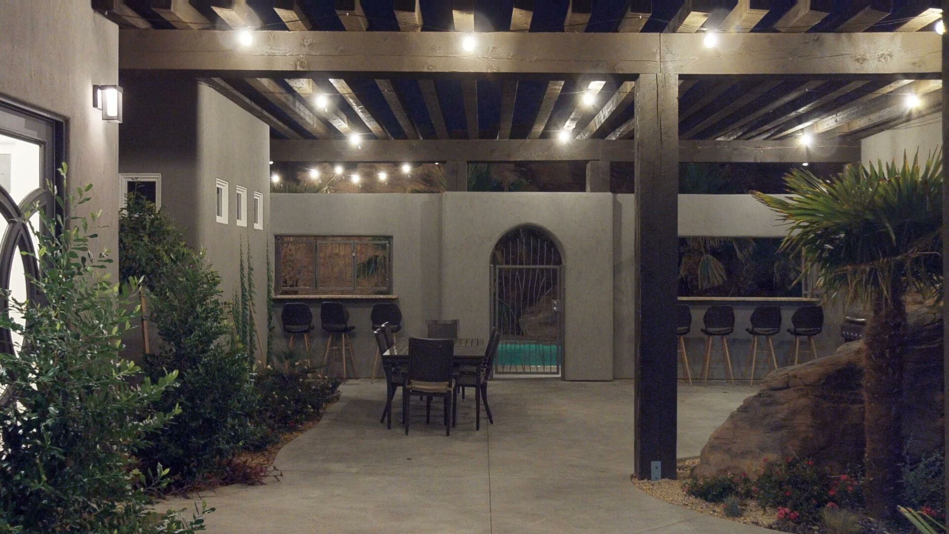 Expansive patio area featuring a slatted wood pergola, built-in bar with seating, a private pool area with an arched entrance, and established landscaping
