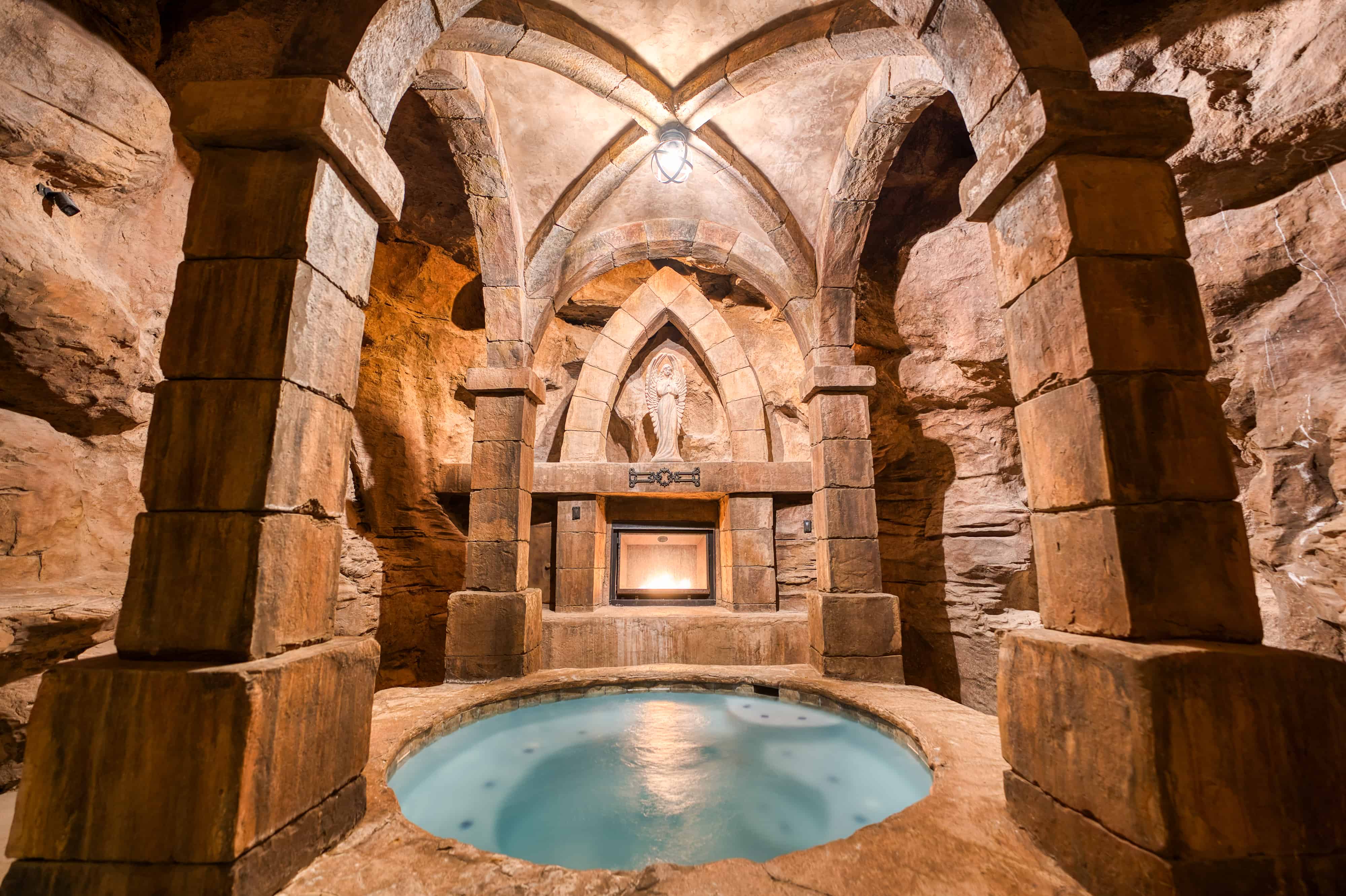 Cavernous grotto featuring a circular spa, stone pillars, vaulted ceilings, integrated fireplace, and carved wall accents