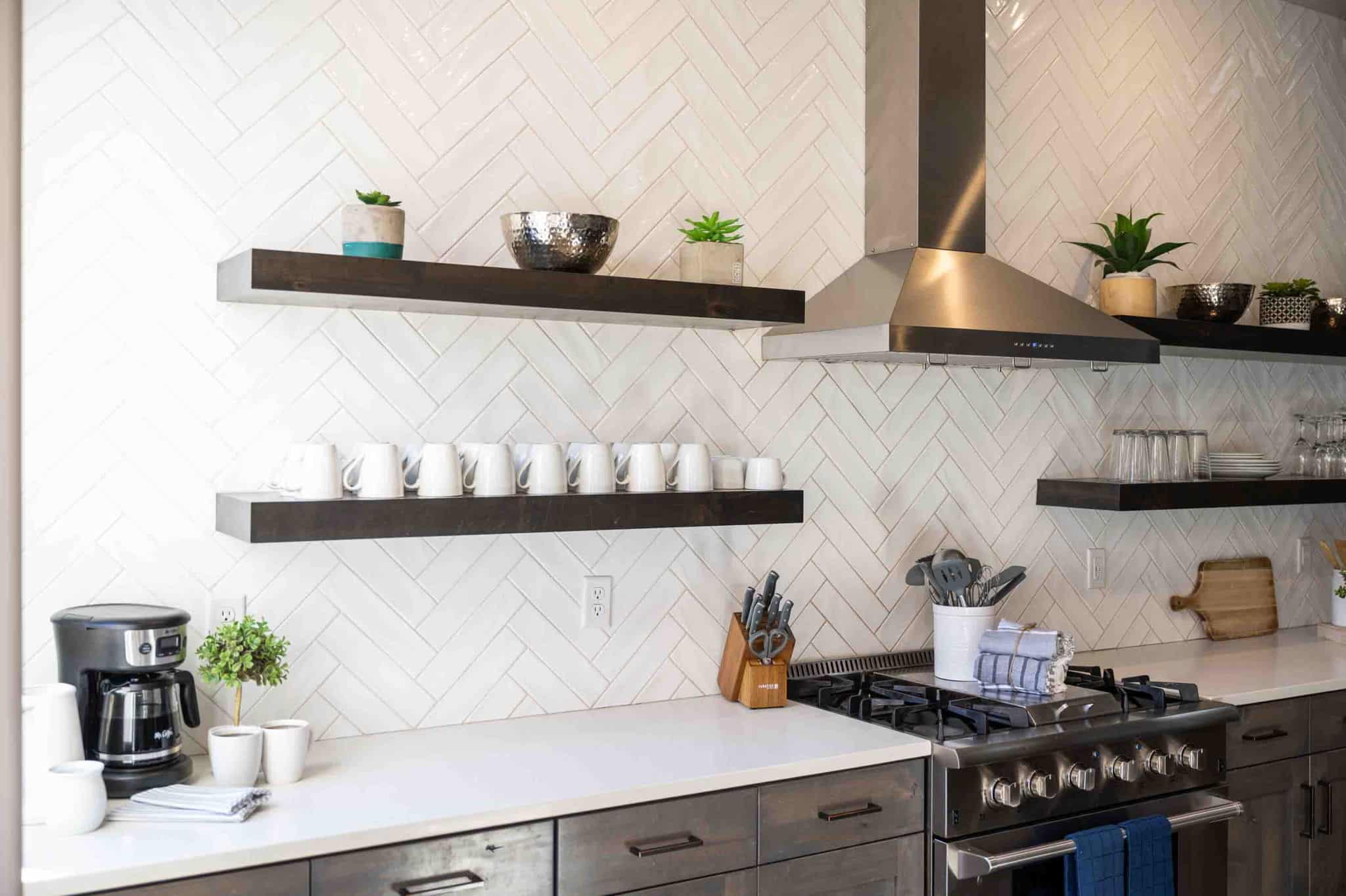 Kitchen featuring white herringbone tile backsplash, stainless steel range hood, stainless steel gas range, dark wood floating shelves, and light-colored countertops