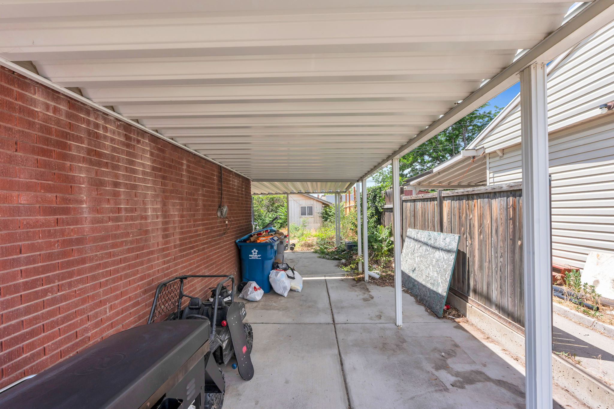Covered concrete walkway featuring a red brick wall, wood panel fencing, and a white metal roof structure