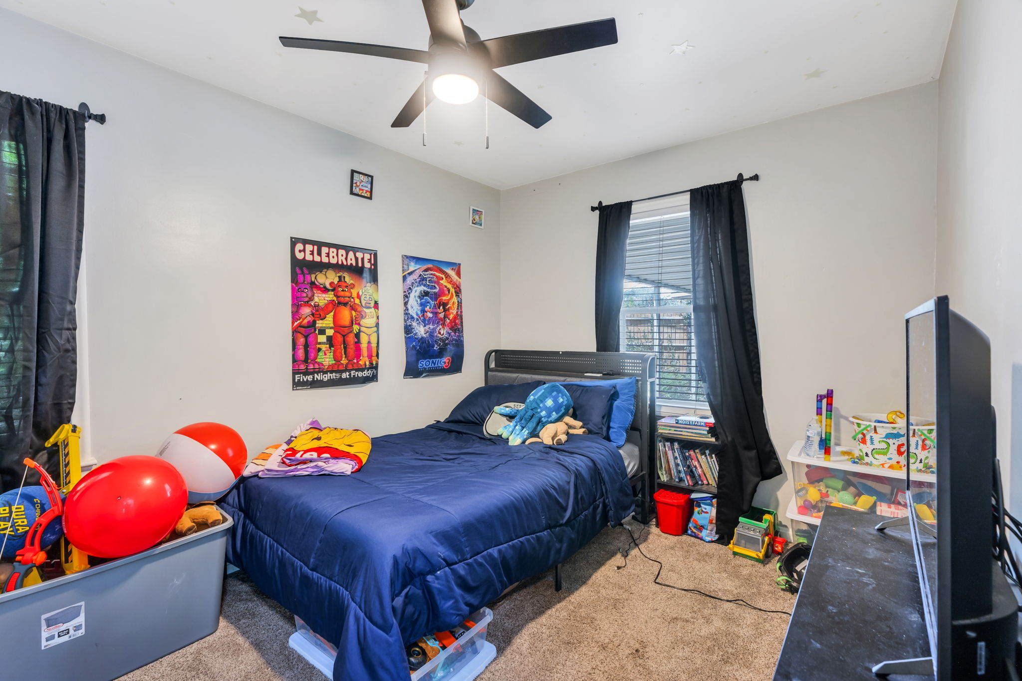 Carpeted bedroom featuring a ceiling fan with integrated lighting, a window with blinds and dark curtains, and light-toned walls