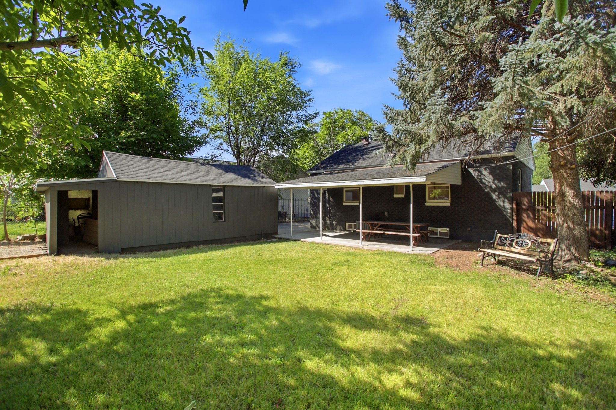 Expansive backyard featuring a manicured lawn, a covered patio, a detached storage shed with an open bay, mature trees, and a privacy fence