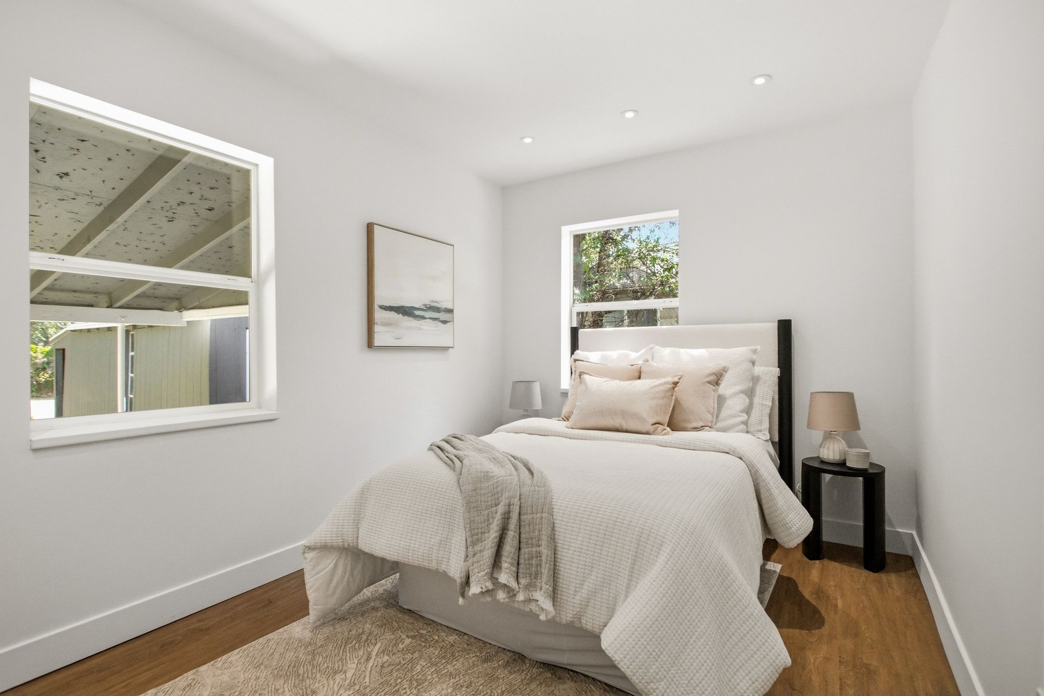 Bright bedroom featuring wood-finish flooring, white baseboards, recessed ceiling lighting, and two windows providing natural light