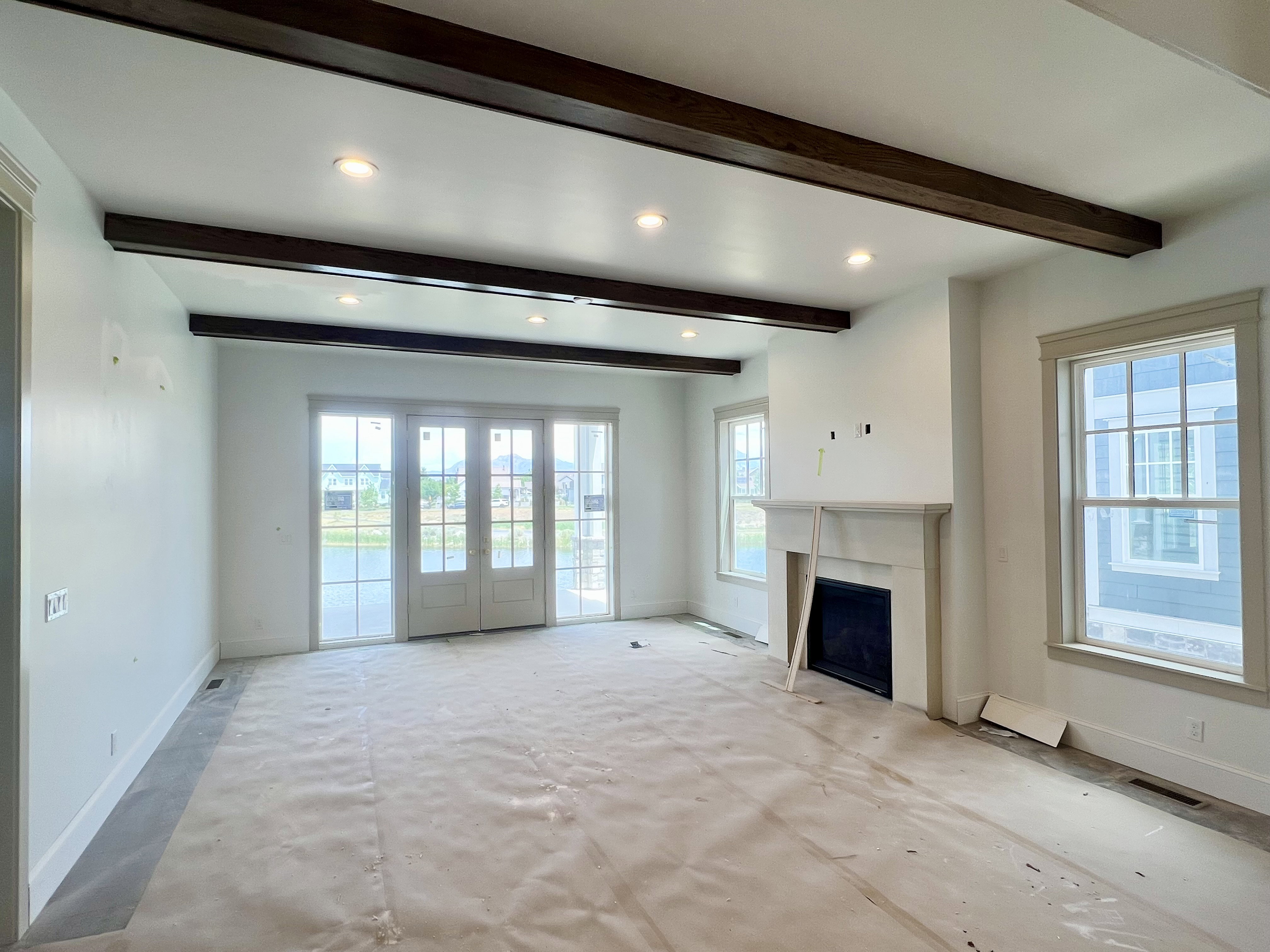 Living area featuring exposed ceiling beams, recessed lighting, double French doors, a prominent fireplace with a substantial mantel, and multiple windows with trim