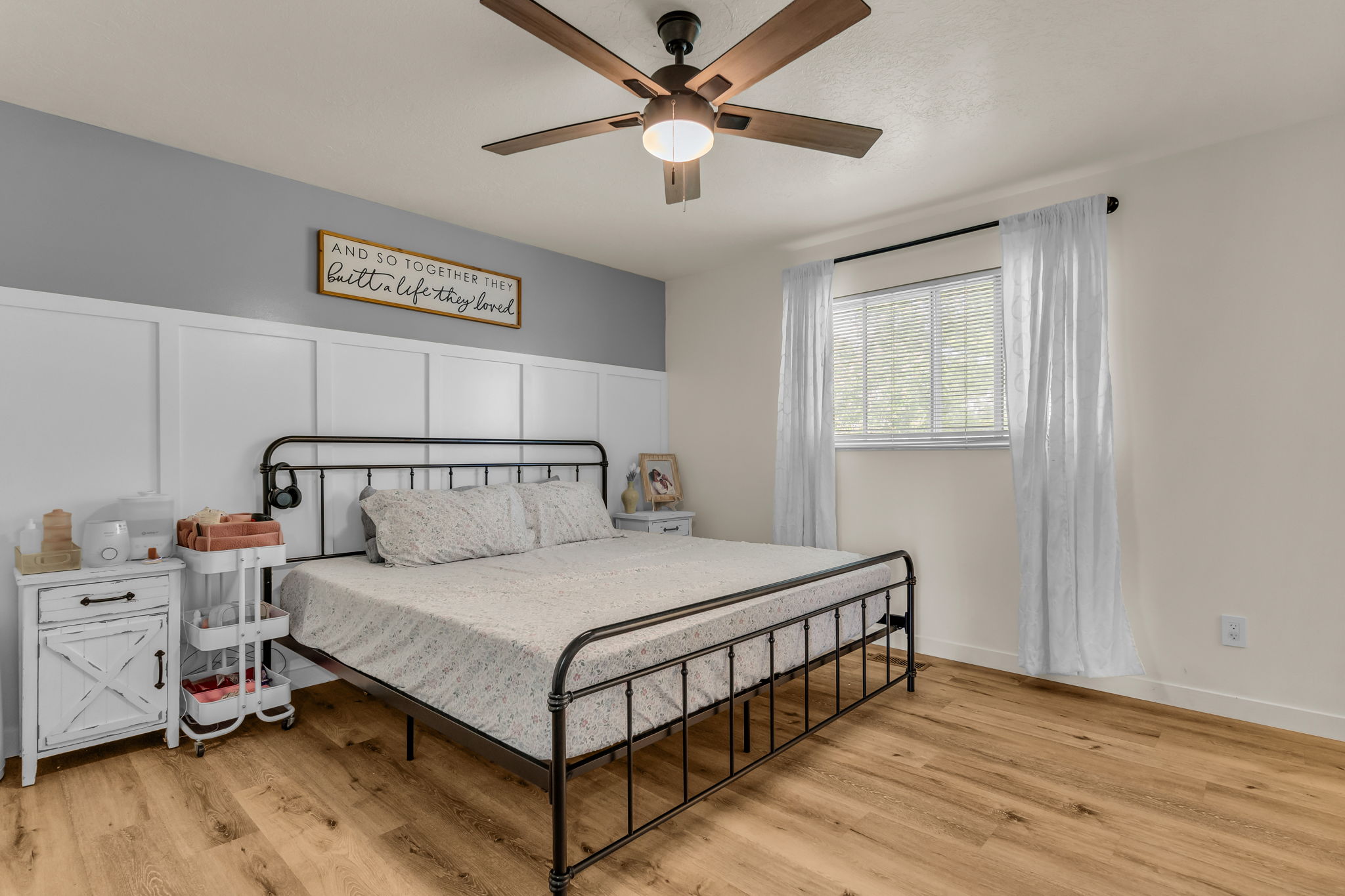 Bedroom with wood-finish flooring, a decorative accent wall featuring wainscoting and a contrasting painted upper portion, a ceiling fan with integrated lighting, and a single window with blinds