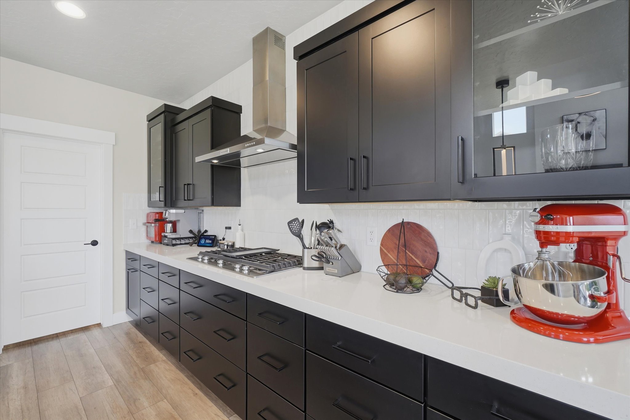 Modern kitchen featuring dark cabinetry with matte black hardware, light-toned countertops, a gas cooktop with stainless steel range hood, a white subway tile backsplash, and wood-finish flooring