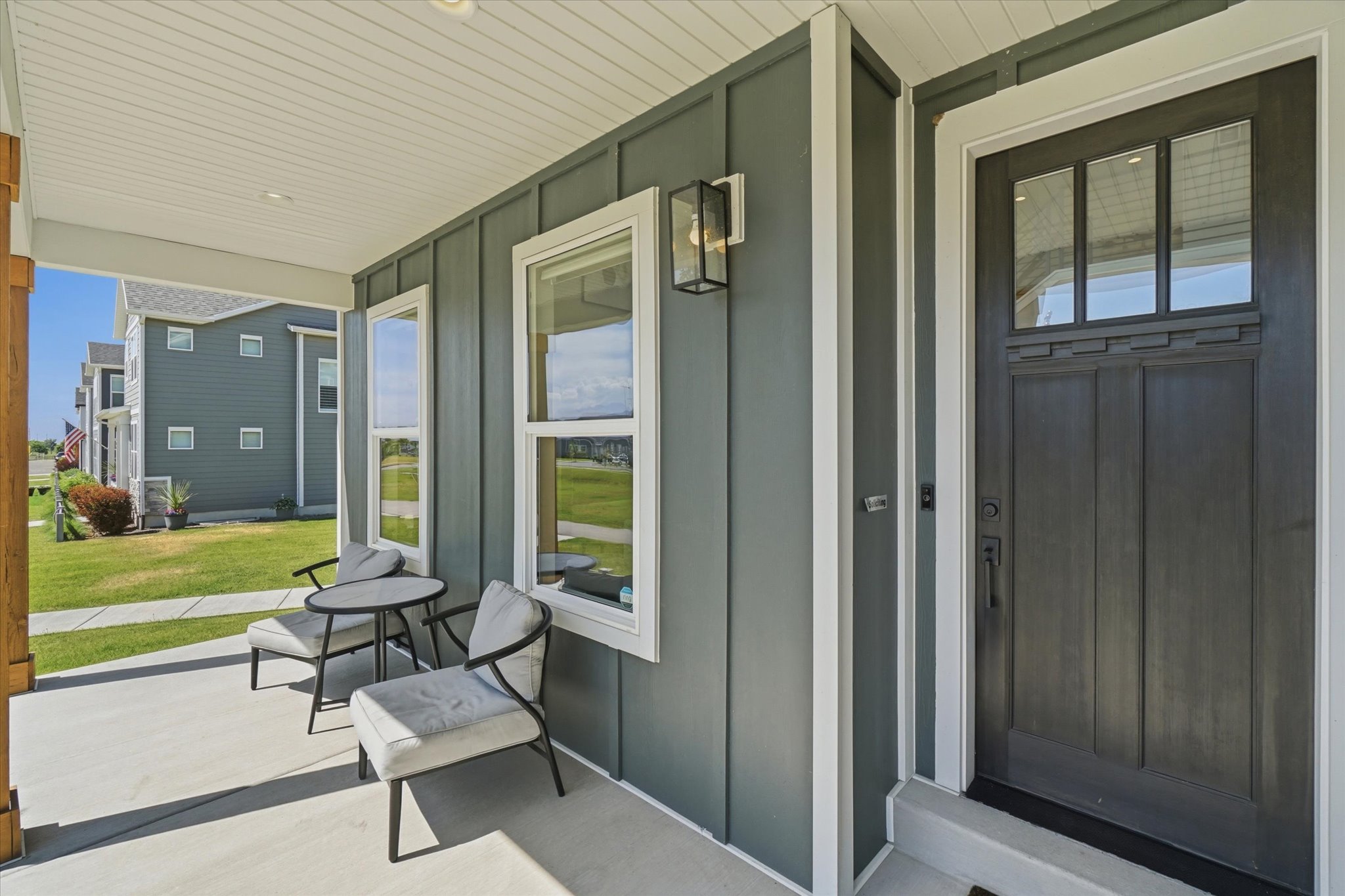Covered concrete porch featuring a substantial dark entry door with upper glass panels, two adjacent vertical windows with white trim, board-and-batten siding, and an exterior wall lantern