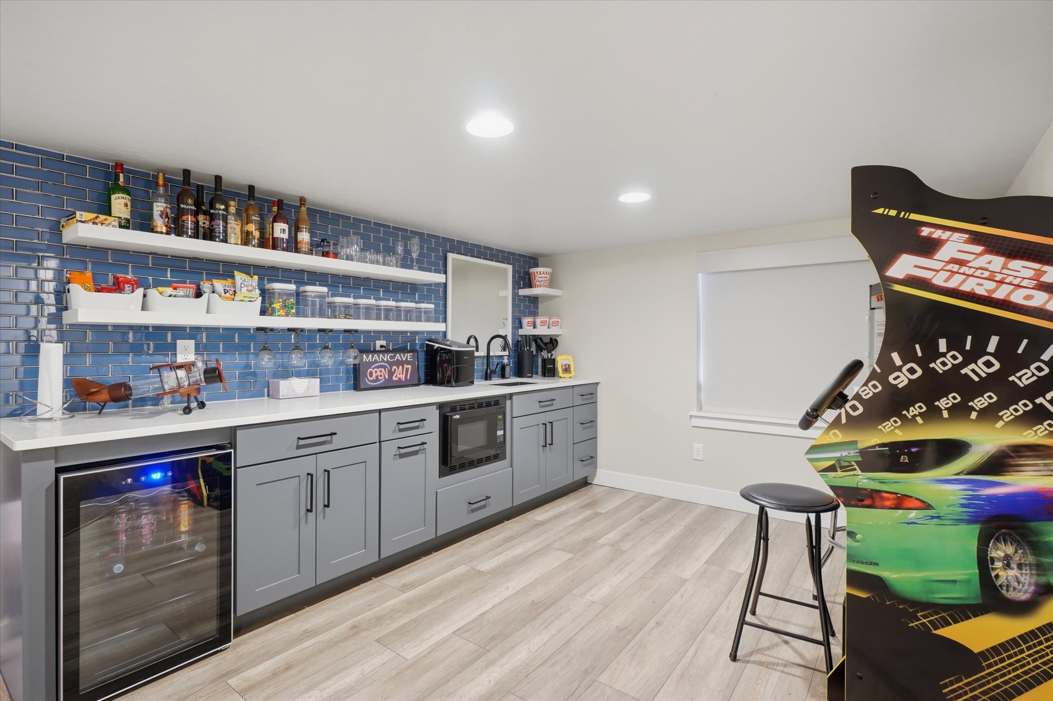 Recreation space featuring a wet bar with a blue subway tile backsplash, white floating shelves, gray cabinetry, and a beverage refrigerator