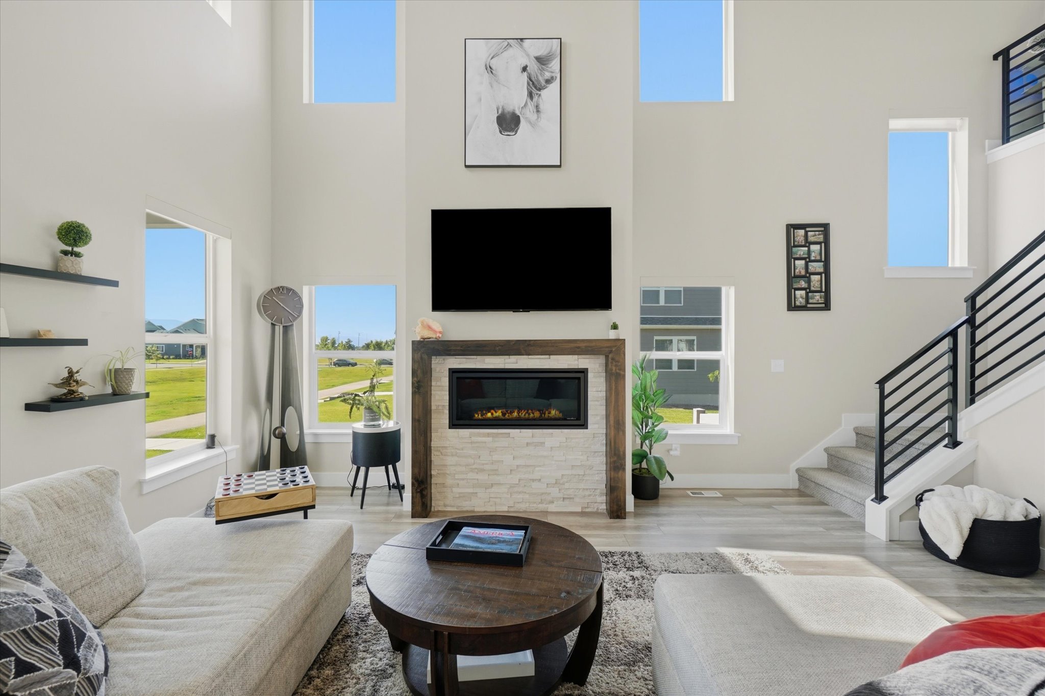 Two-story living area featuring a stacked stone fireplace with a wood-finish mantle, large windows, and an open staircase with black metal railings