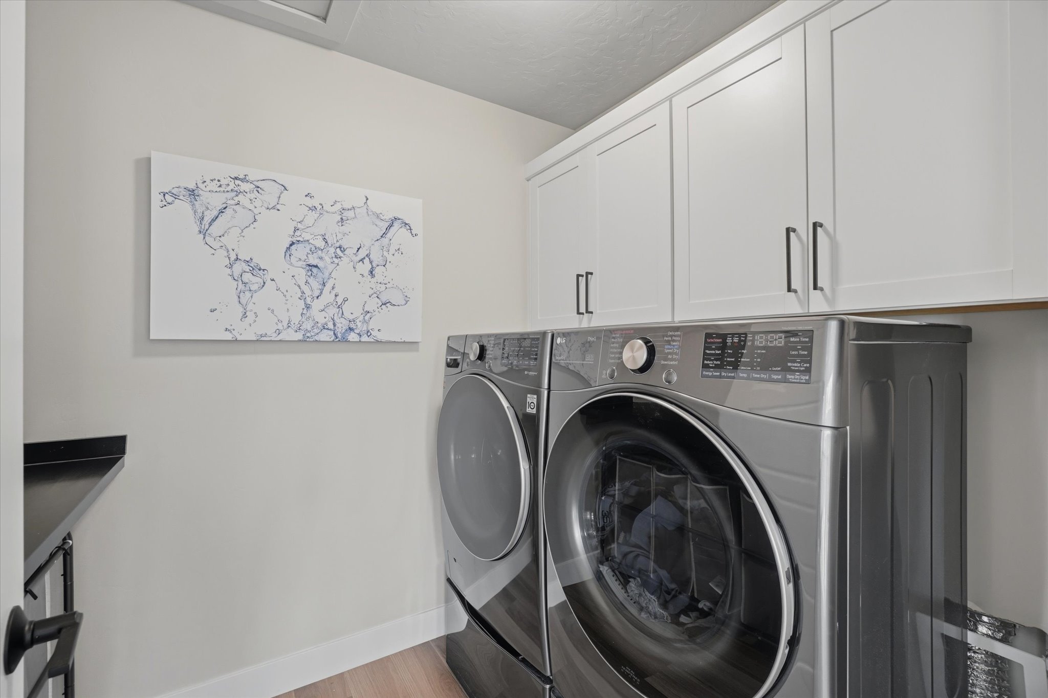 Laundry area featuring light wood-finish flooring, white shaker-style cabinetry with dark hardware, and a dark countertop