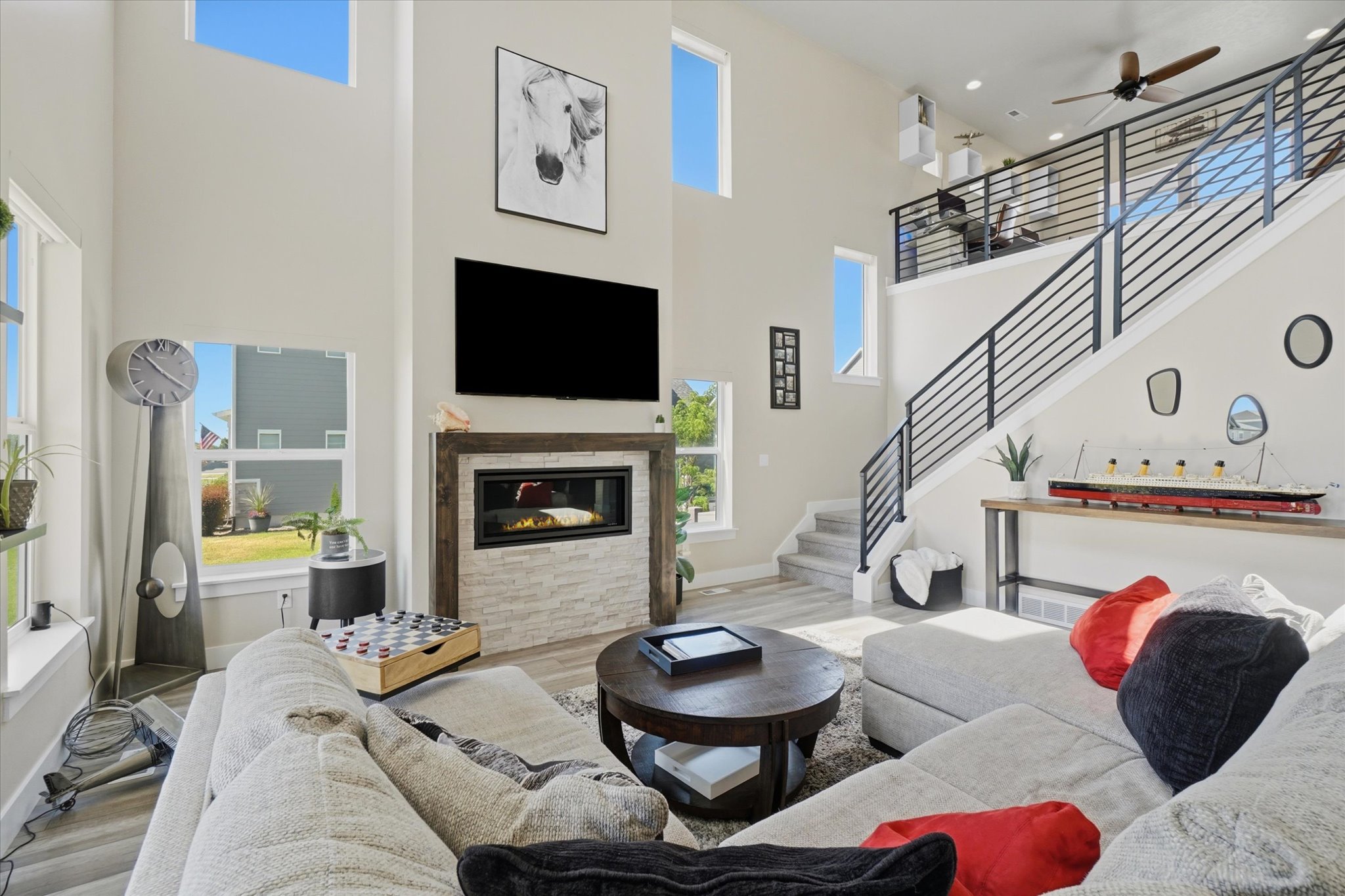 Spacious living area featuring a two-story ceiling, multiple windows, a modern fireplace with stone surround, a contemporary staircase with metal railings, and wide plank flooring