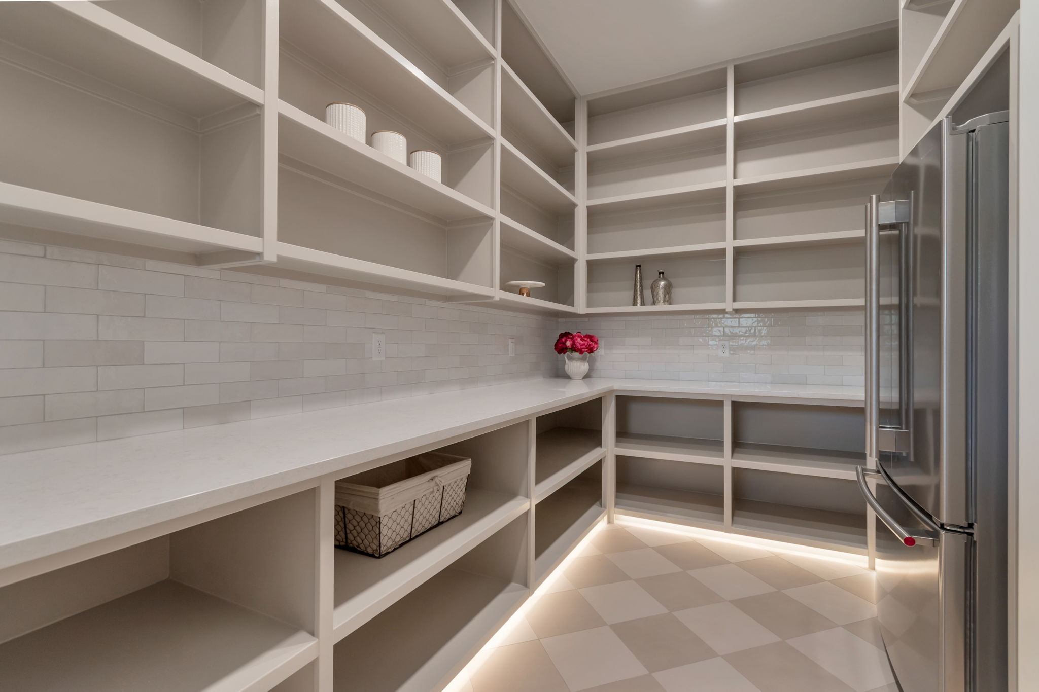 Spacious pantry featuring extensive built-in shelving, ample countertop space, a subway tile backsplash, stainless steel refrigerator, and checkerboard floor tile with under-shelf lighting