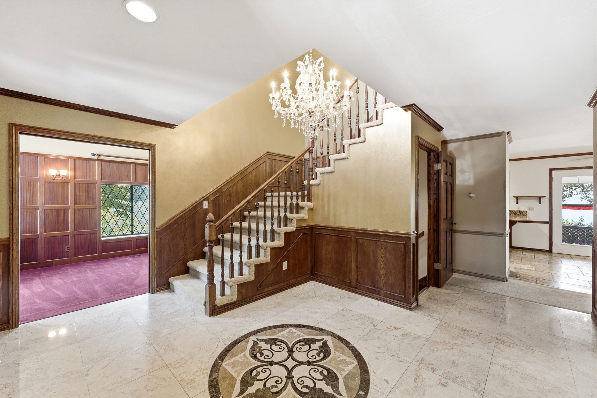 Grand foyer featuring a crystal chandelier, wood-paneled staircase. View of living room and walks into kitchen