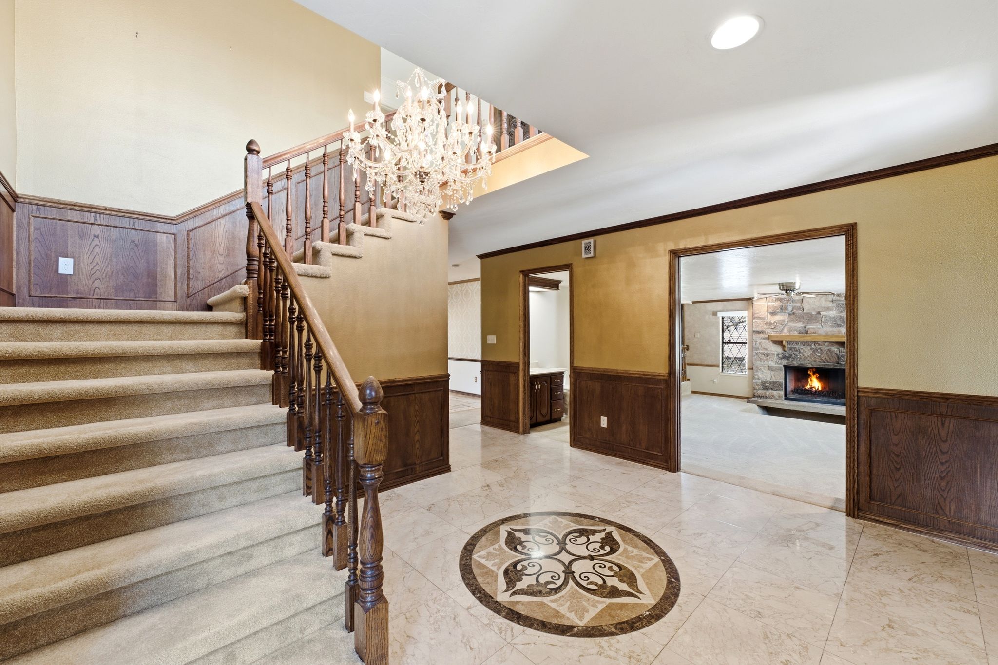 Grand entry foyer featuring light-toned tile flooring with an intricate medallion inlay, a carpeted staircase with wood railing, and a multi-tiered crystal chandelier