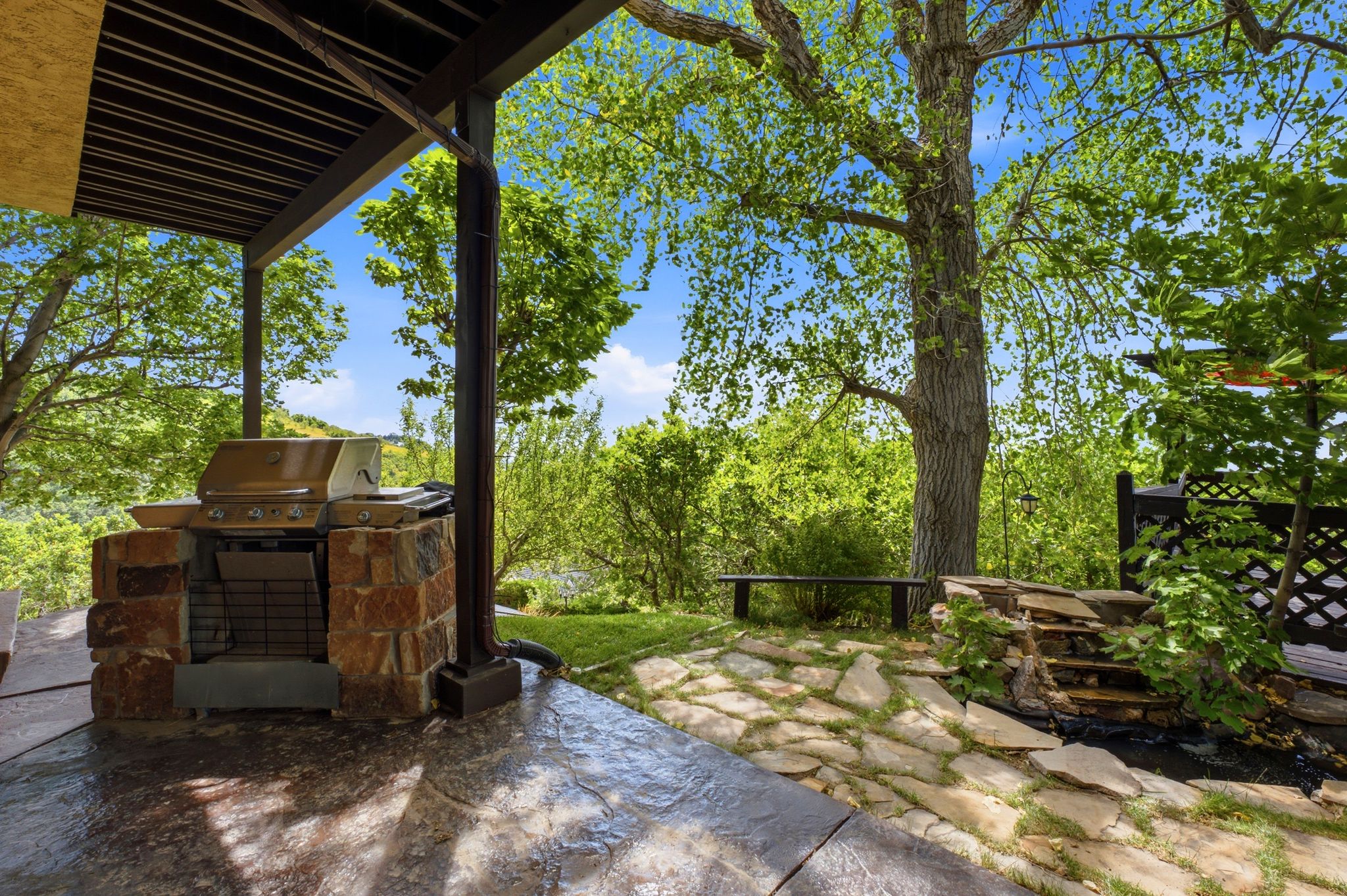 Covered outdoor patio featuring a built-in stone barbecue, stamped concrete flooring, flagstone pathway, and mature trees