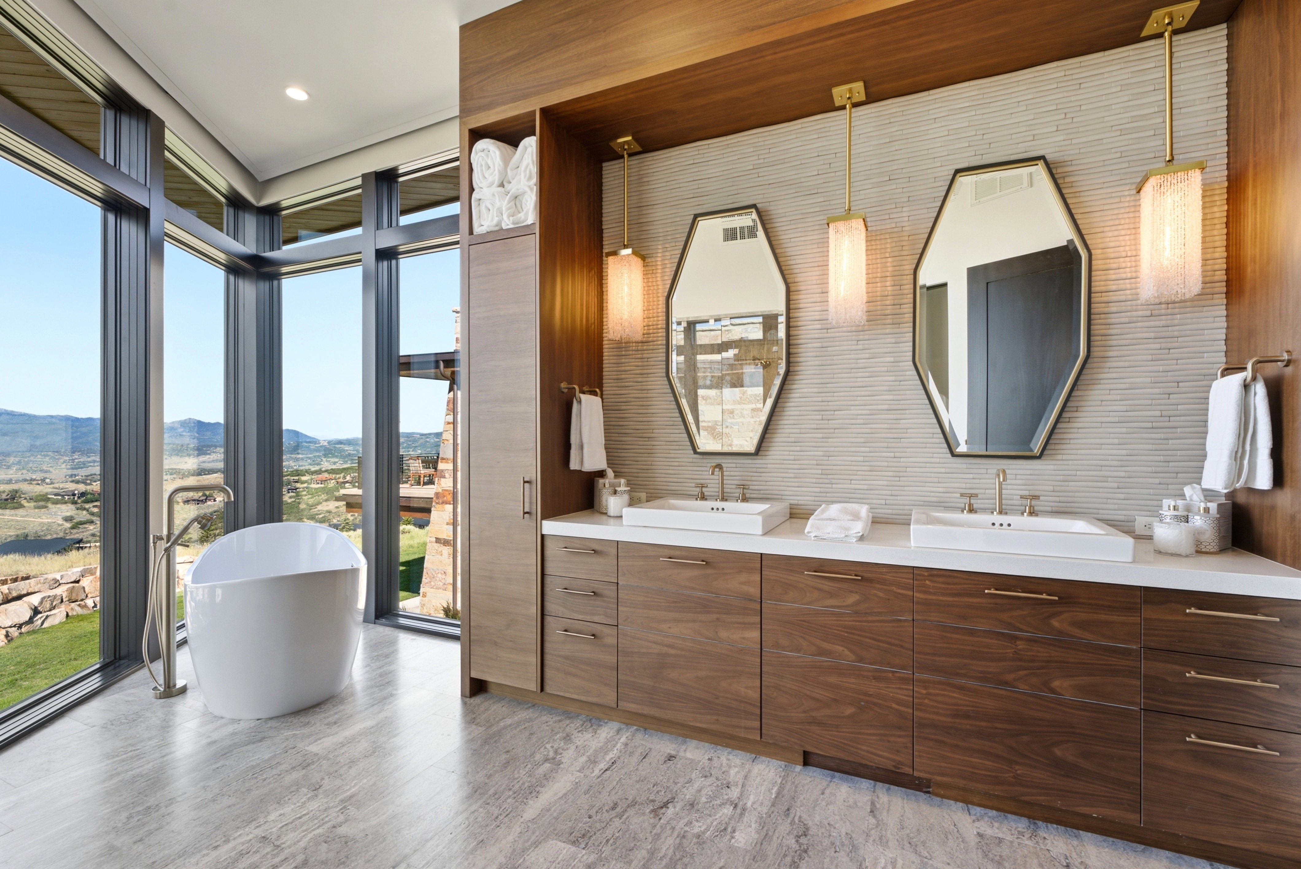 Contemporary bathroom featuring a freestanding tub, floor-to-ceiling windows, a dual vanity with wood-finish cabinetry, a textured stone accent wall, and geometric mirrors
