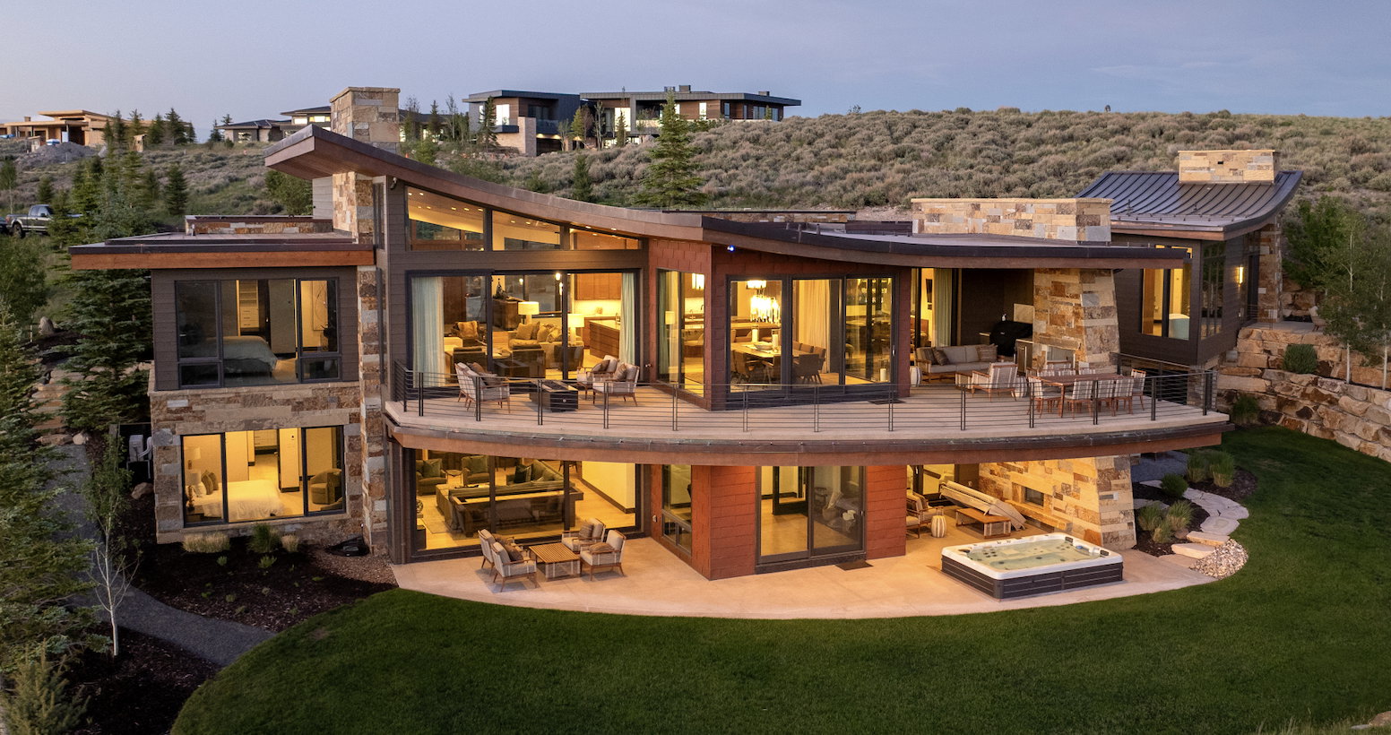 Contemporary multi-level residence featuring expansive window walls, stone and wood-finish siding, multiple outdoor entertaining areas, and a lower-level patio with an integrated spa