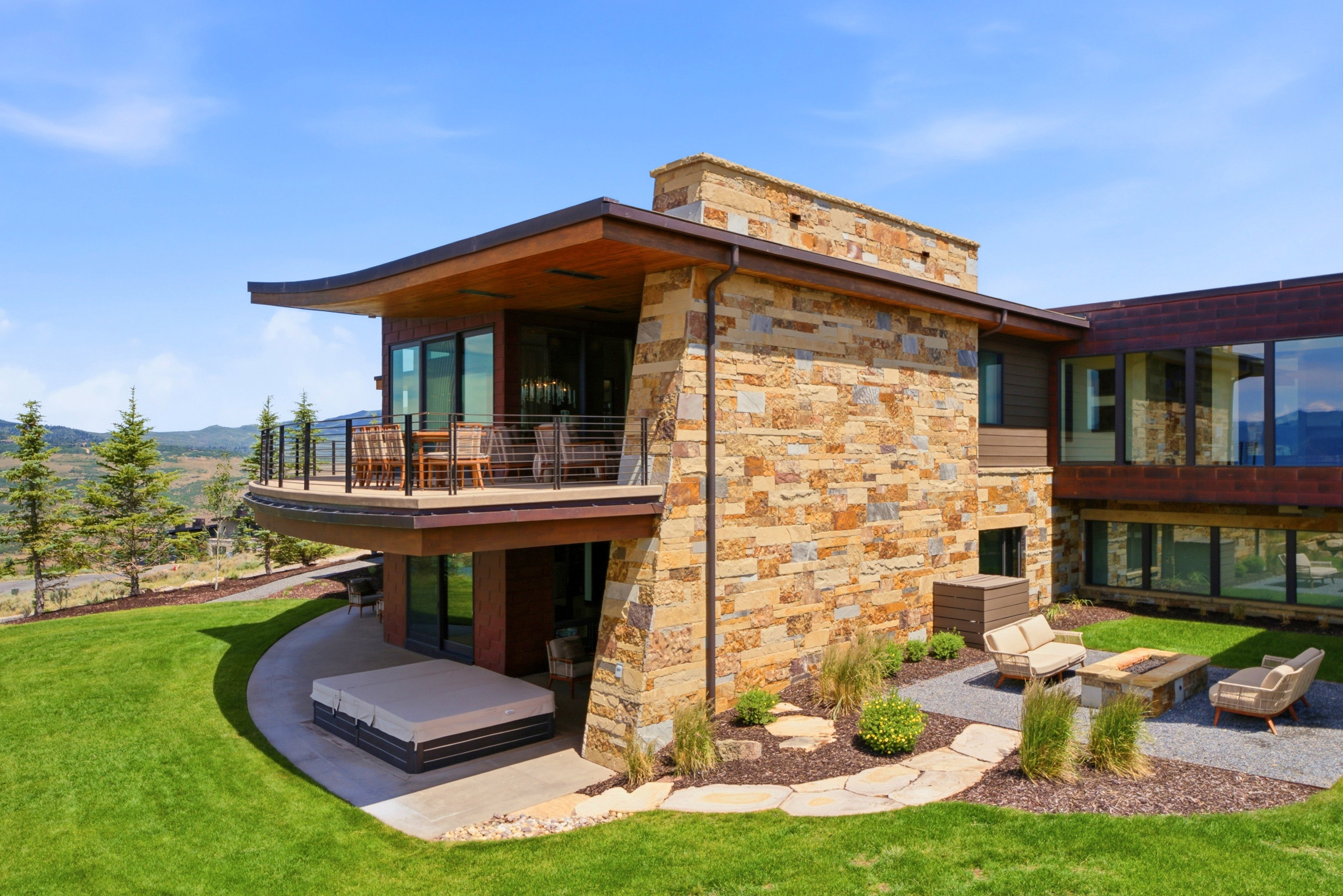 Contemporary architecture featuring extensive natural stone cladding, a curved upper-level balcony with metal railings, and a lower-level patio with a spa