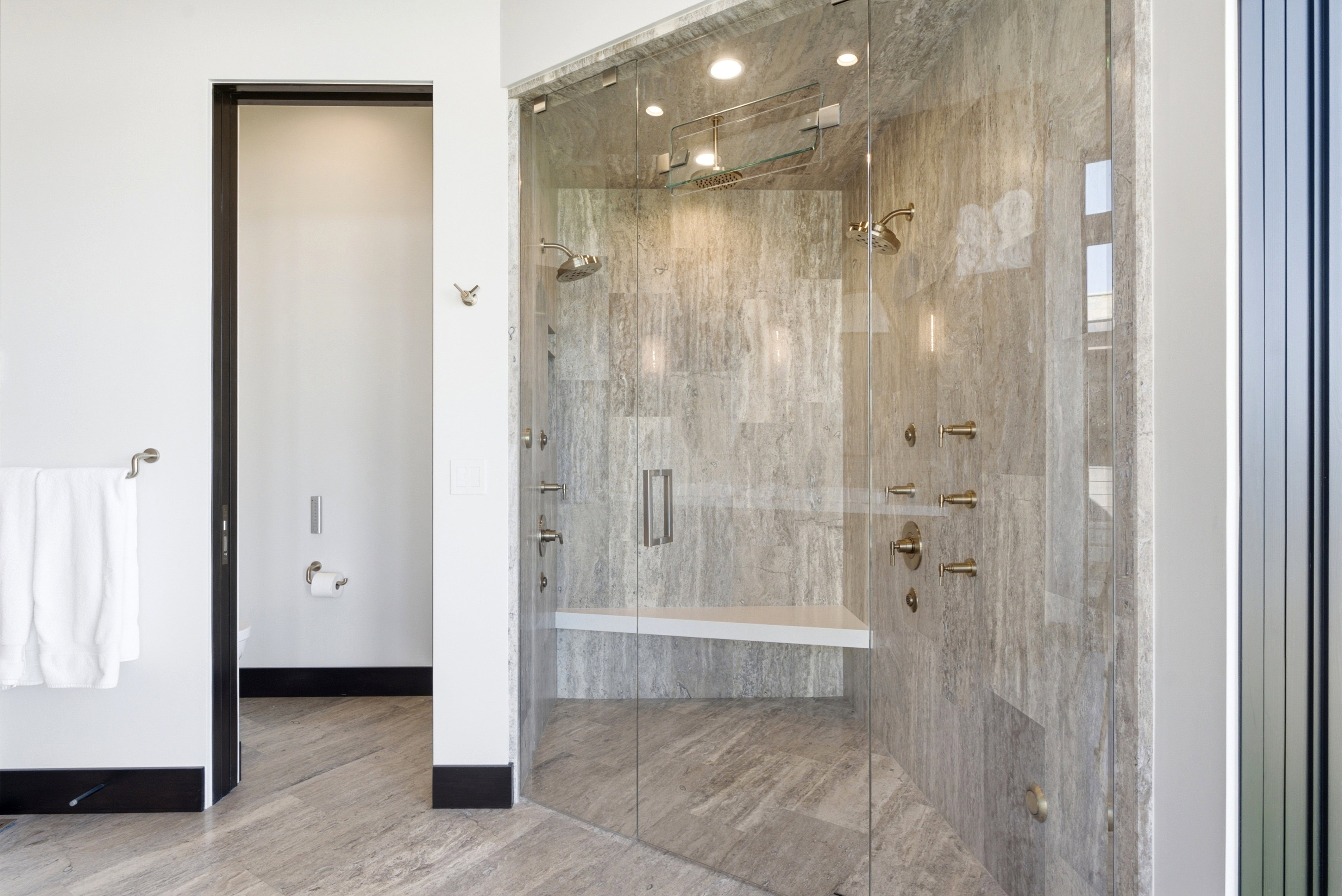 Spacious shower enclosure with floor-to-ceiling glass, featuring large format stone tile, a built-in bench, multiple showerheads, and integrated lighting