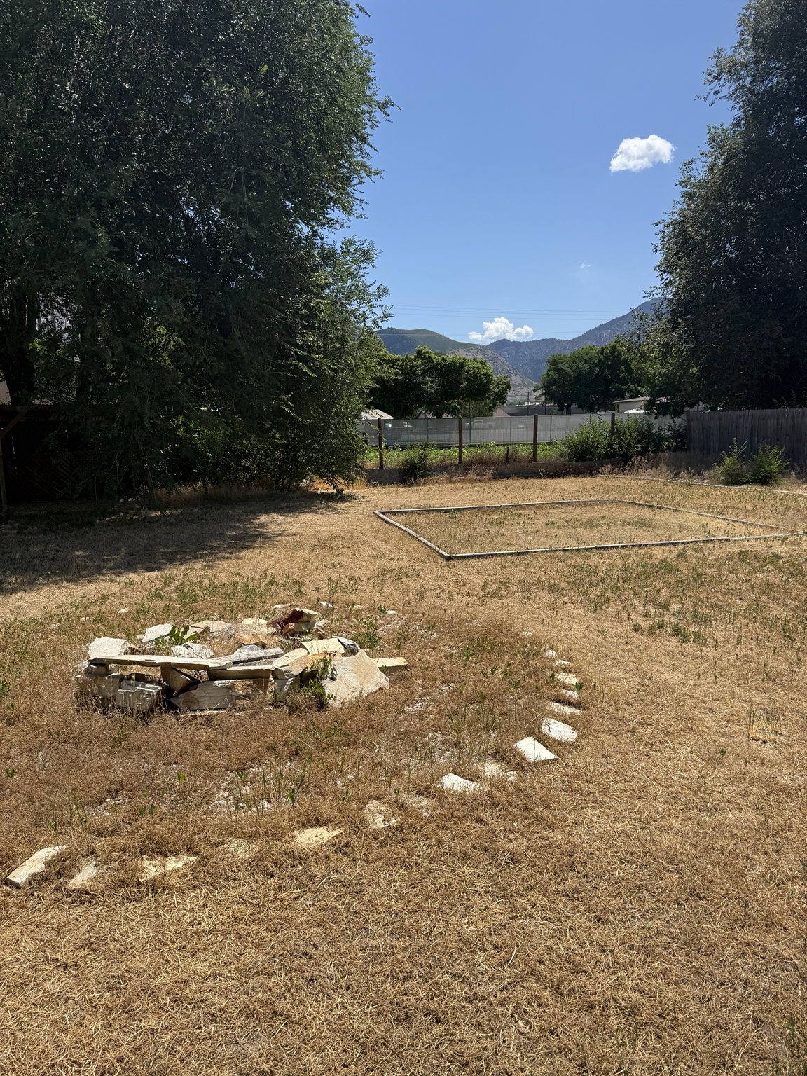 Expansive backyard featuring a stone-lined fire pit, a defined garden bed, mature perimeter trees, and mountain scenery
