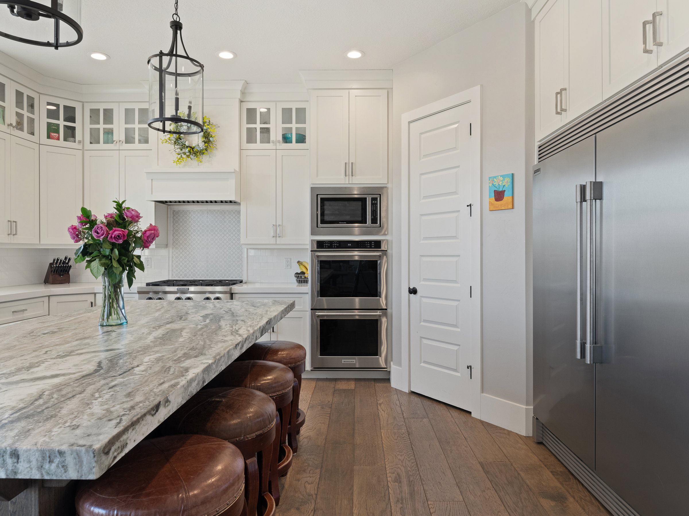 Spacious kitchen featuring a large island with a substantial stone countertop, wood-finish flooring, white cabinetry, stainless steel appliances, and a built-in double oven