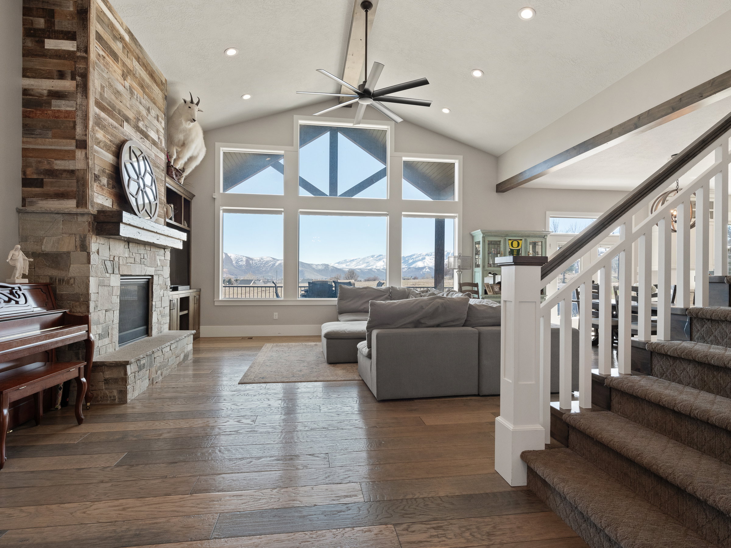 Vaulted ceiling great room featuring a stone fireplace with wood-finish mantel, multi-pane windows showcasing mountain scenery, wood-finish flooring, and a contemporary ceiling fan