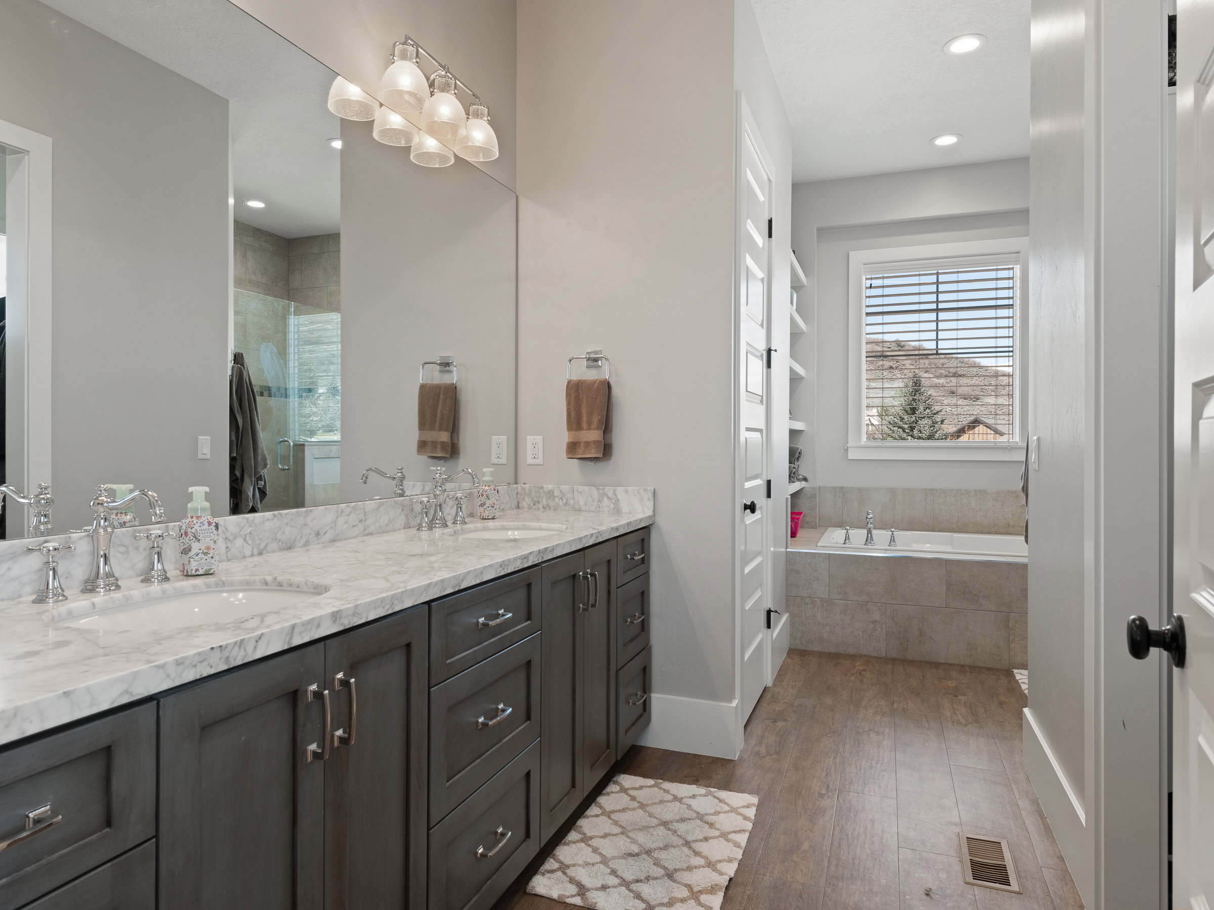 Bathroom featuring a double vanity with stone countertops and undermount sinks, wood-finish flooring, built-in shelving, and a tiled soaking tub