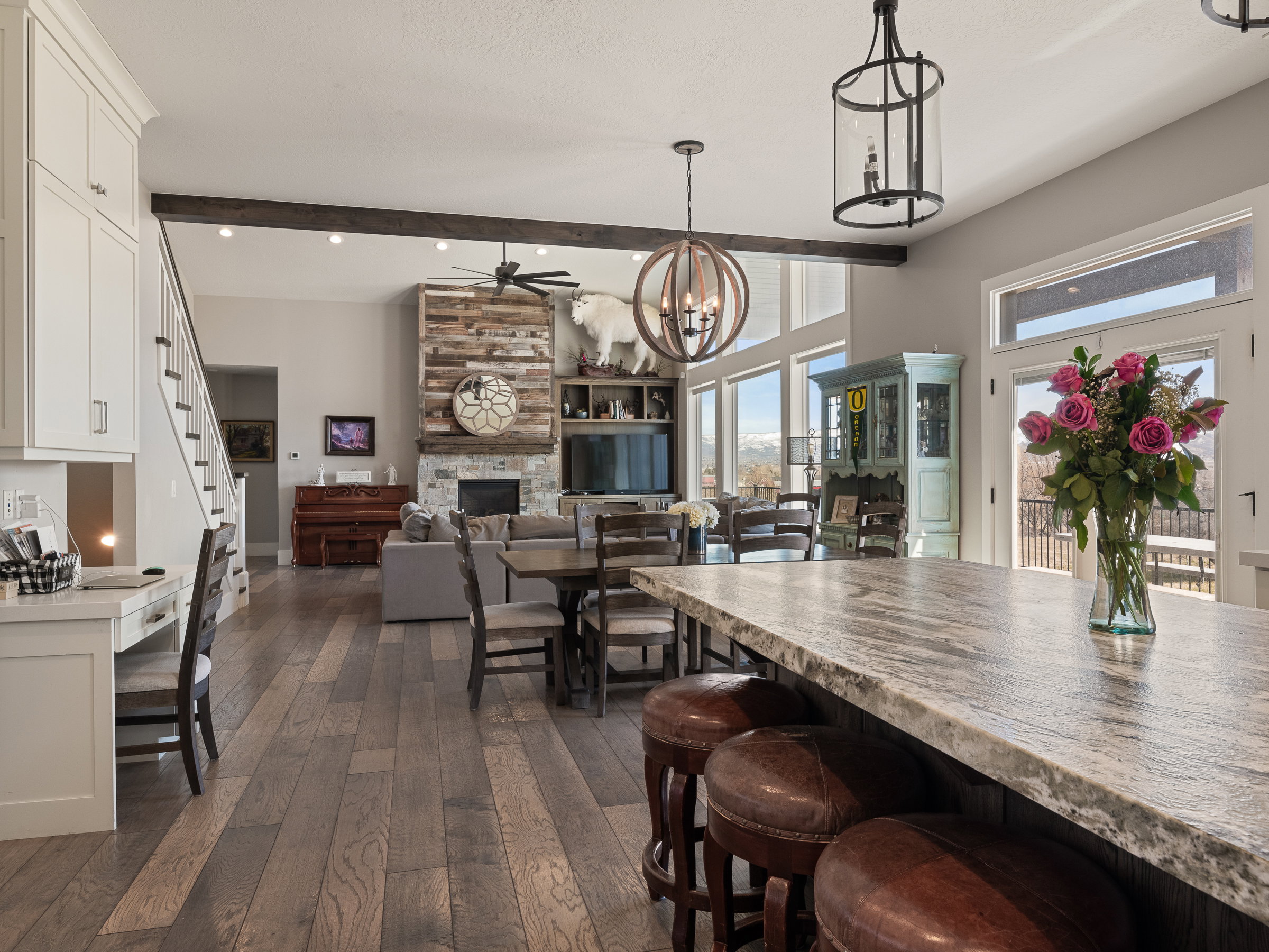 Spacious open-concept interior featuring wood-finish flooring throughout, a stone-faced fireplace with wood paneling, a kitchen island with a stone countertop, a built-in desk area, and large windows