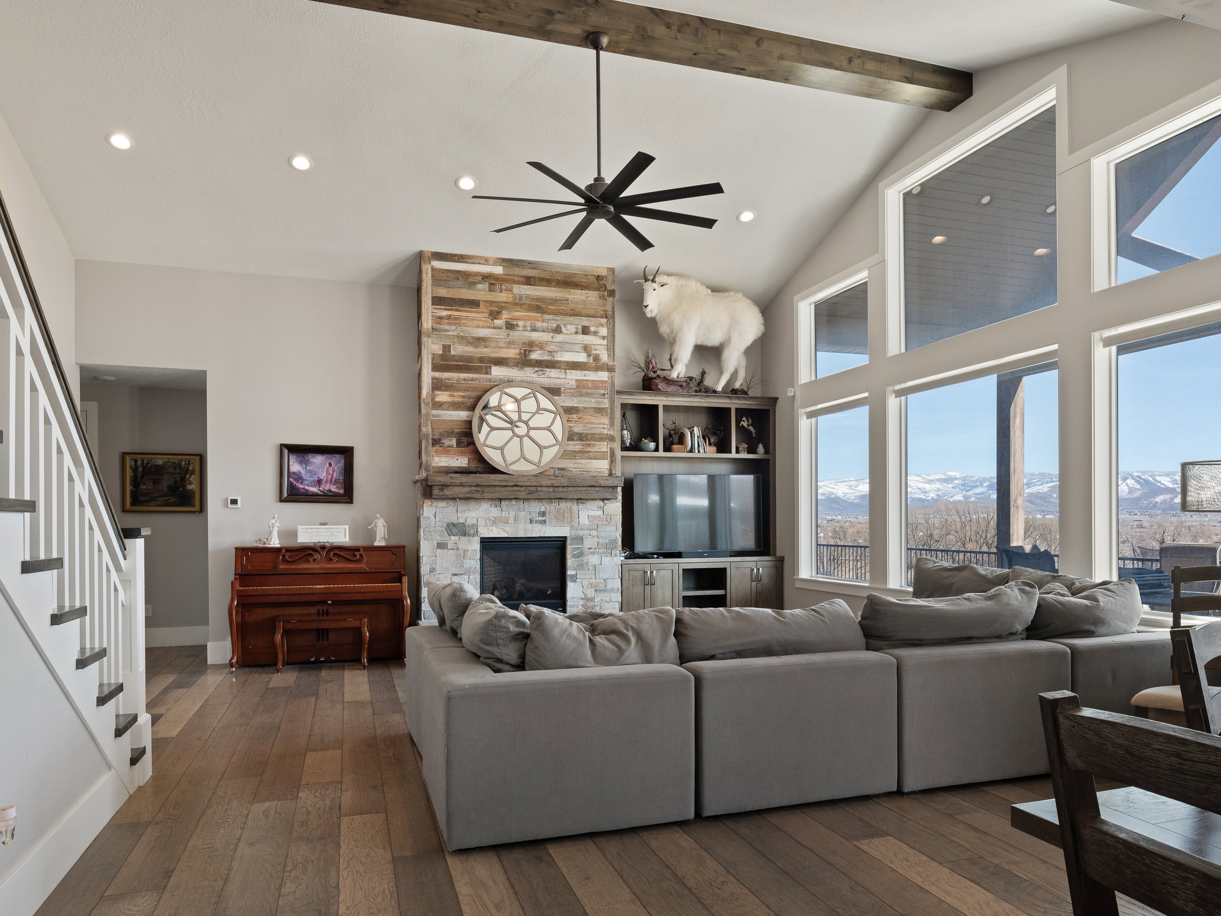 Vaulted ceiling great room with wood-finish flooring, a stone and wood fireplace, an exposed wood beam, large picture windows, and a black ceiling fan