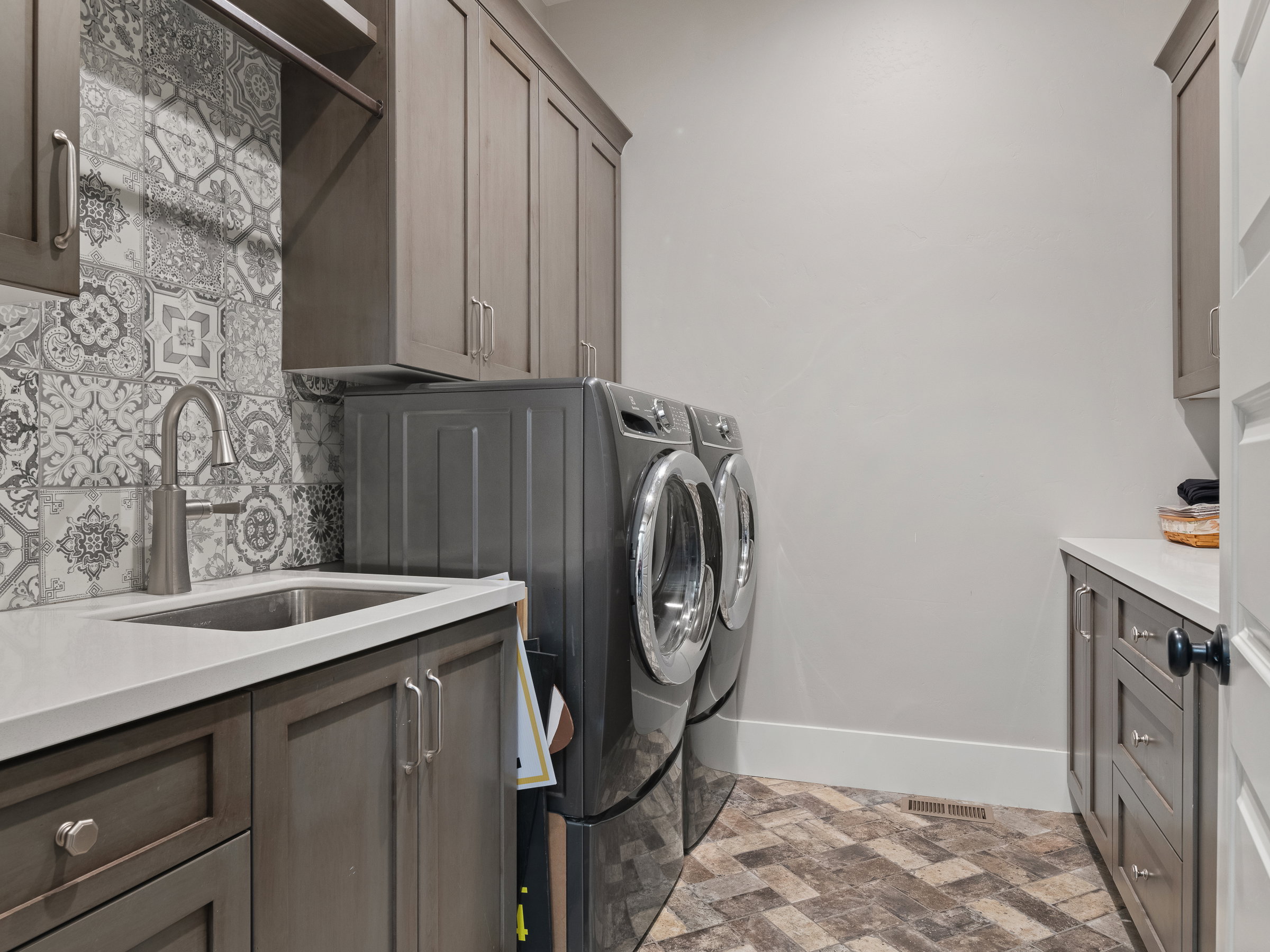 Utility space featuring patterned tile flooring, a stainless steel sink with a high-arc faucet, quartz countertops, extensive cabinetry, and decorative backsplash tiling