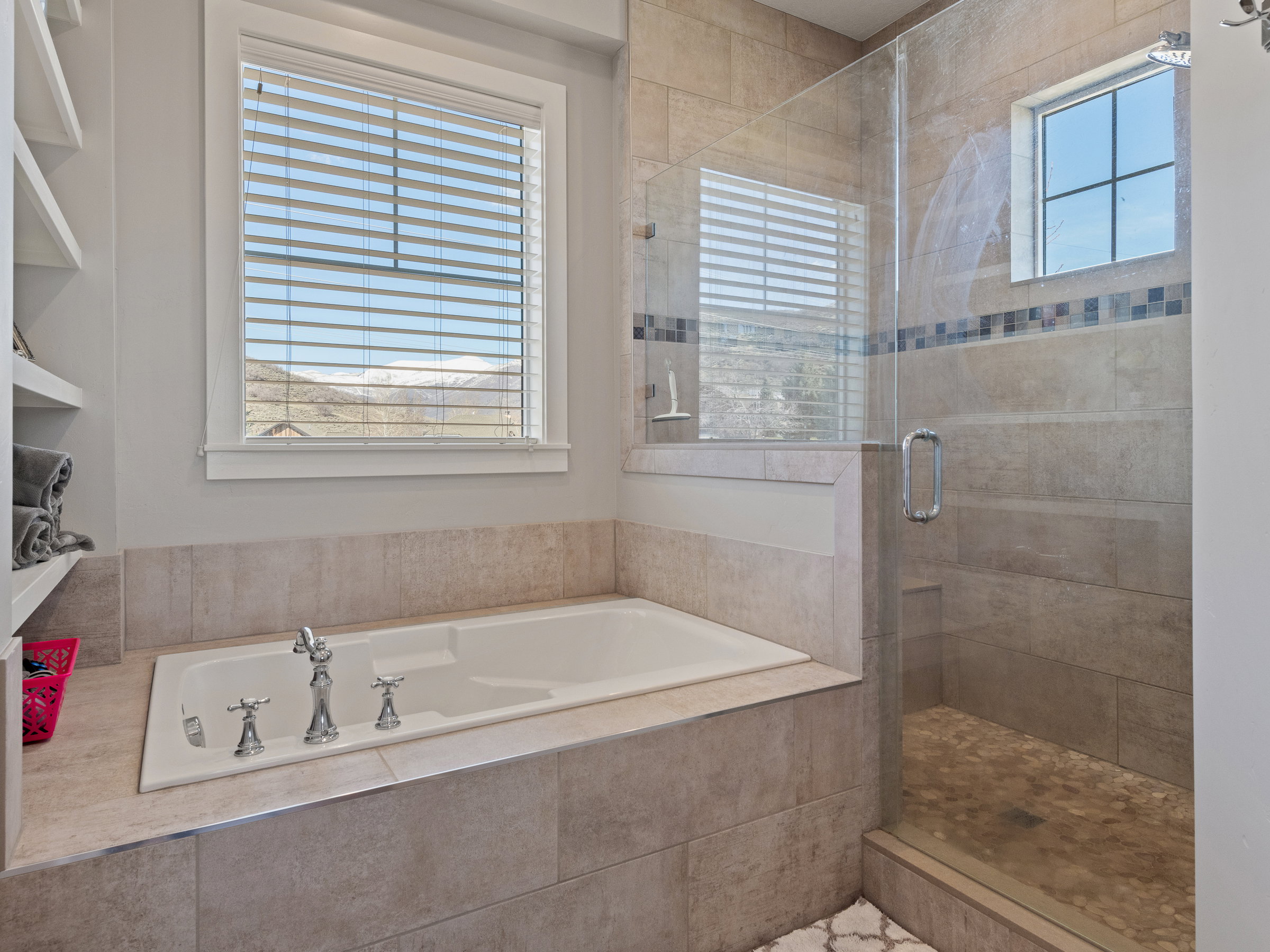 Built-in soaking tub with chrome fixtures, glass-enclosed shower with pebble floor, tile surround, accent tile band, and a window for natural light