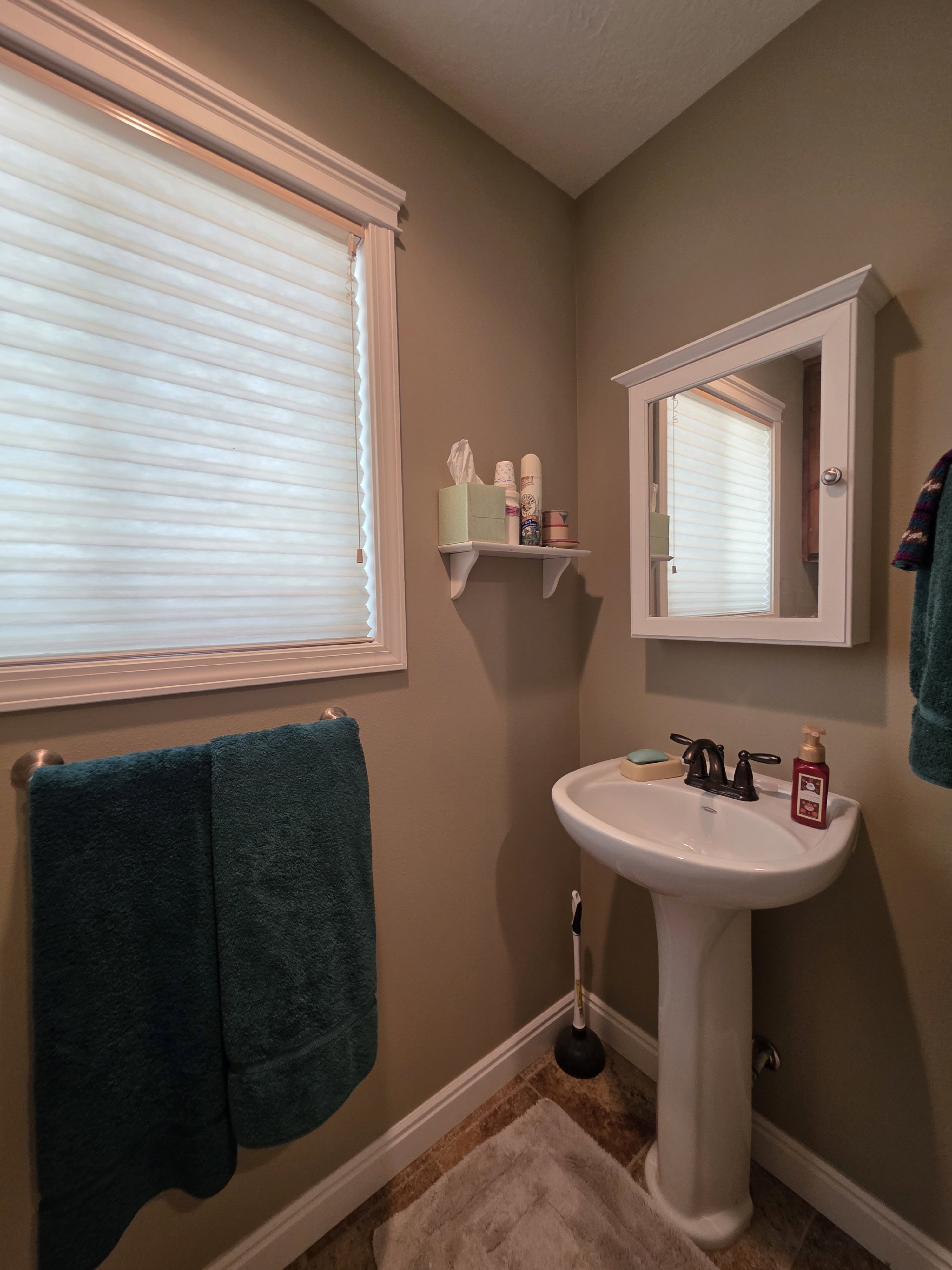 Pedestal sink with dark-finished faucet, framed window with pleated shade, recessed medicine cabinet, wall-mounted shelf, and wood-finish flooring