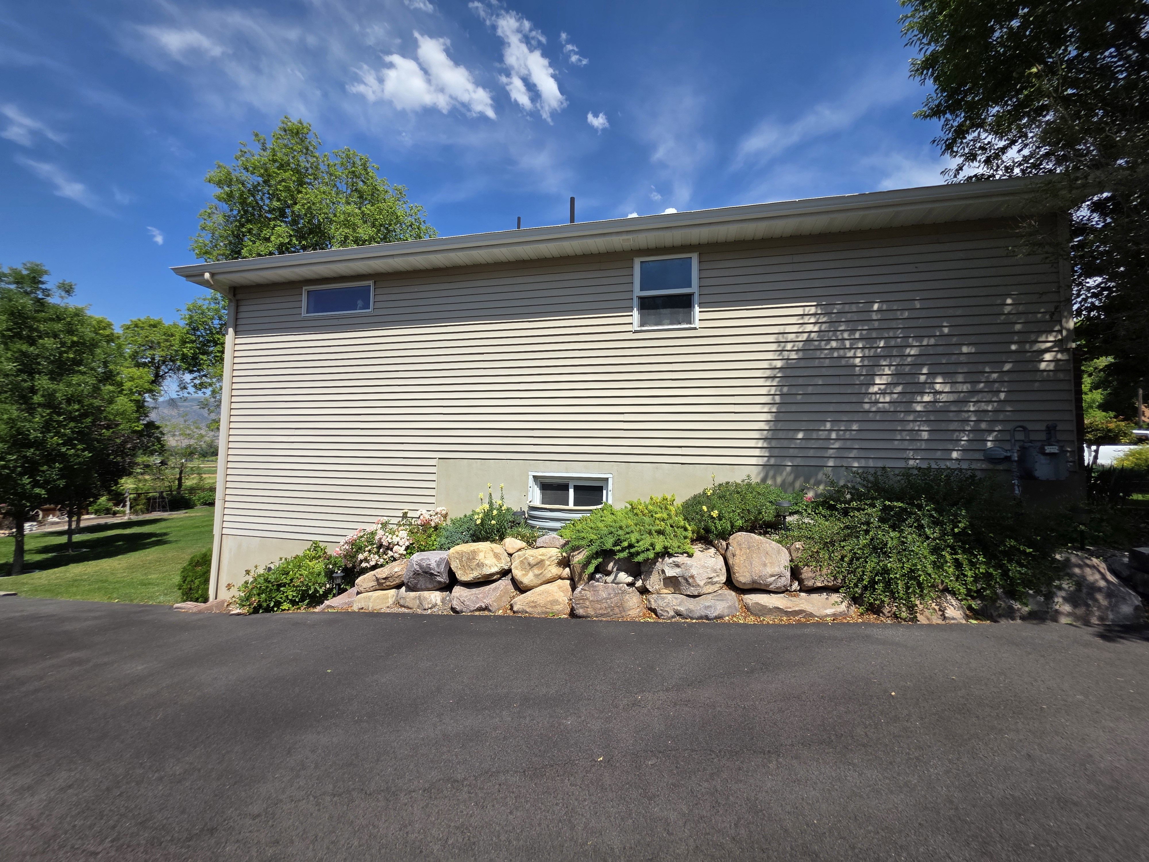 Tan horizontal siding exterior with a rock retaining wall and mature landscaping