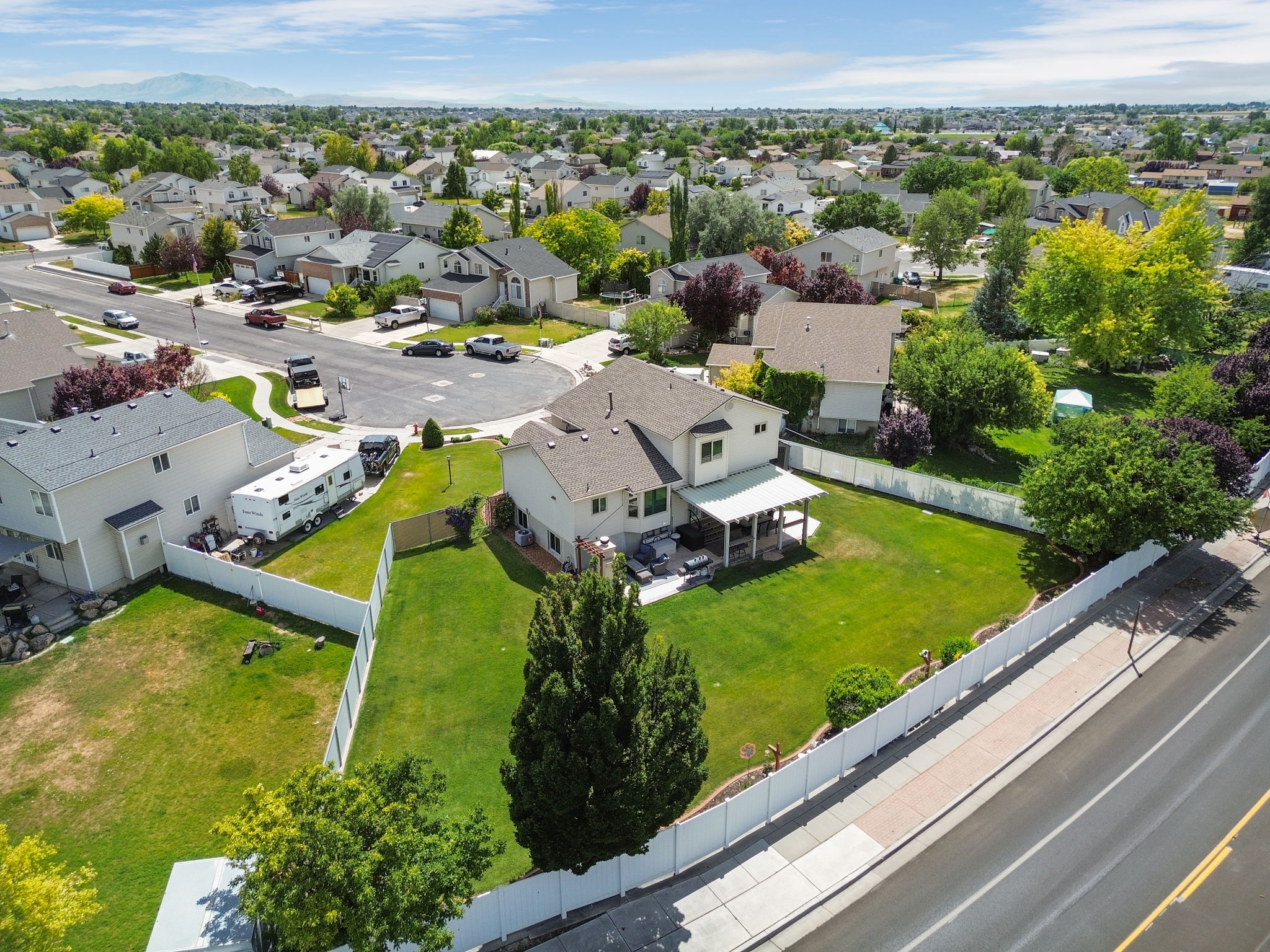 Expansive fenced backyard featuring a manicured lawn, established mature trees, a covered patio, and a two-story residence with a light-colored exterior