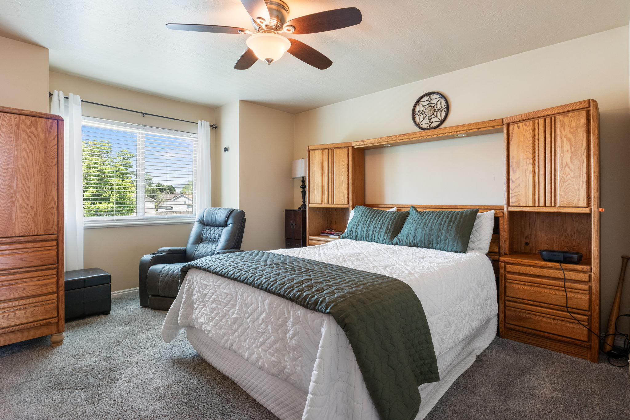 Primary bedroom featuring a large window with blinds, ceiling fan with integrated lighting, and a built-in wood headboard unit with shelving and storage