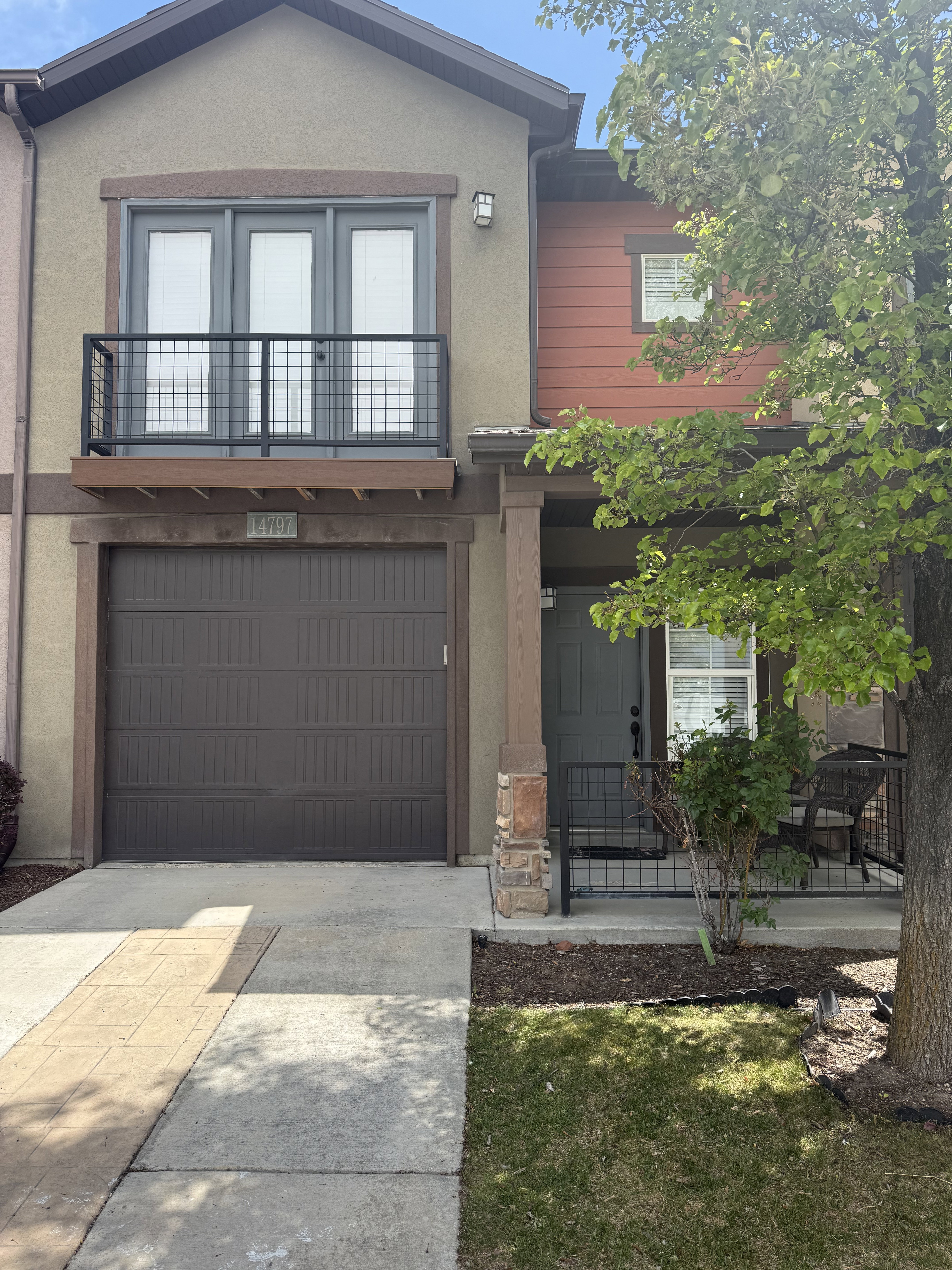 Contemporary townhouse exterior featuring a two-car garage, upper-level balcony with metal railing, stone-clad porch column, and a distinct red siding accent