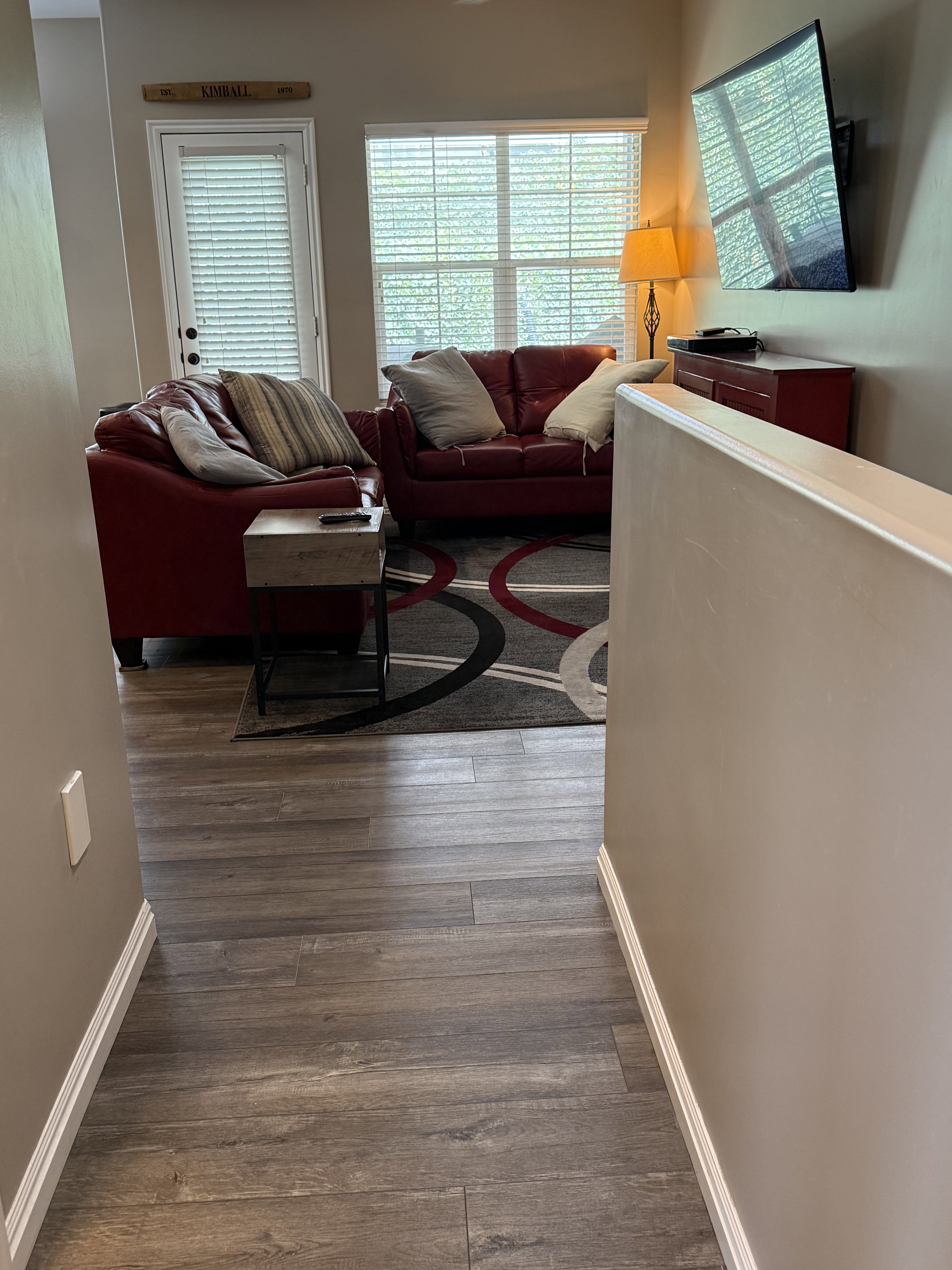 Living area featuring wood-finish flooring, a contemporary area rug, a large window with blinds, and a built-in wall-mounted television