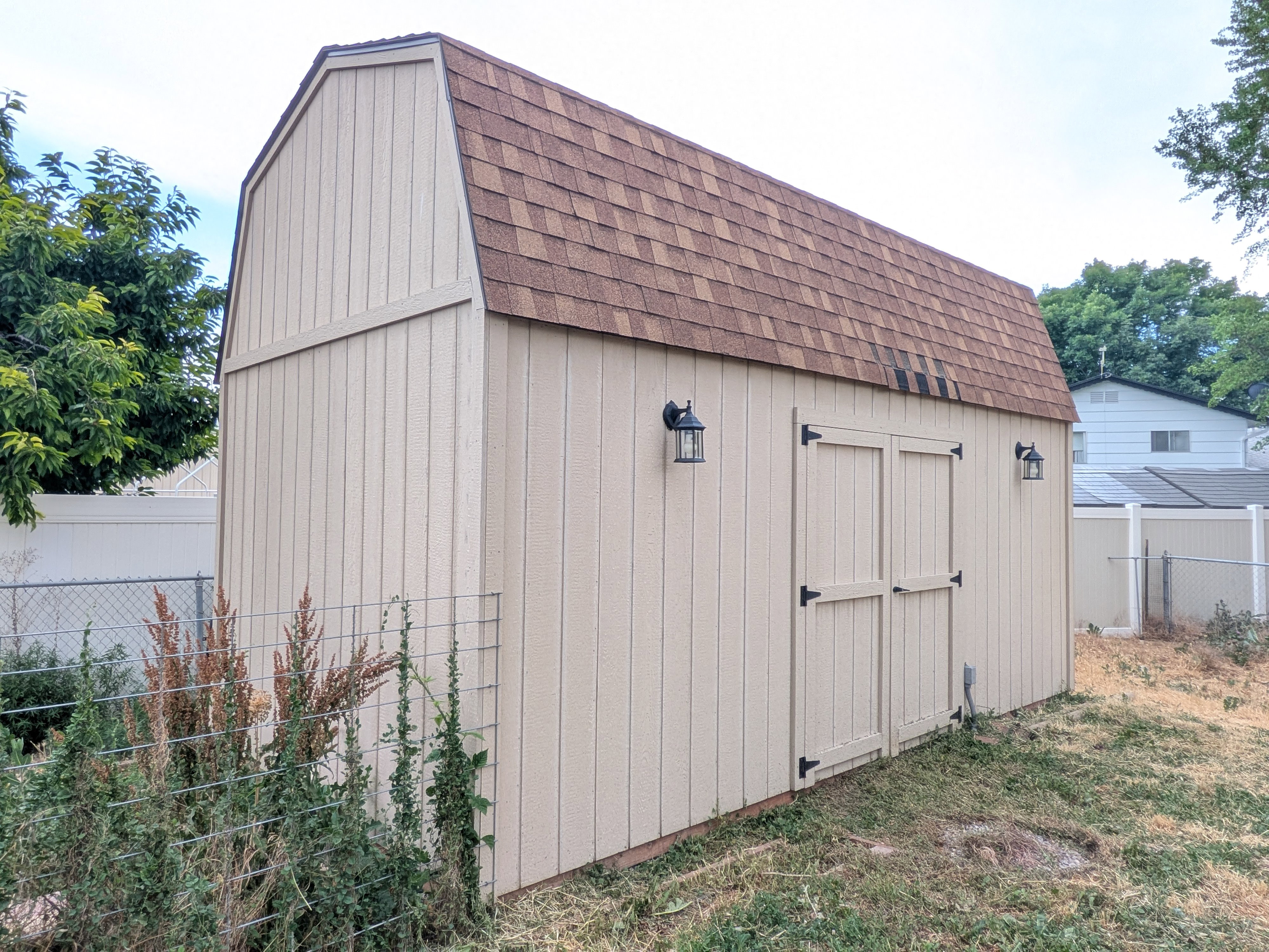 Gambrel-style shed with beige vertical siding and a brown shingle roof