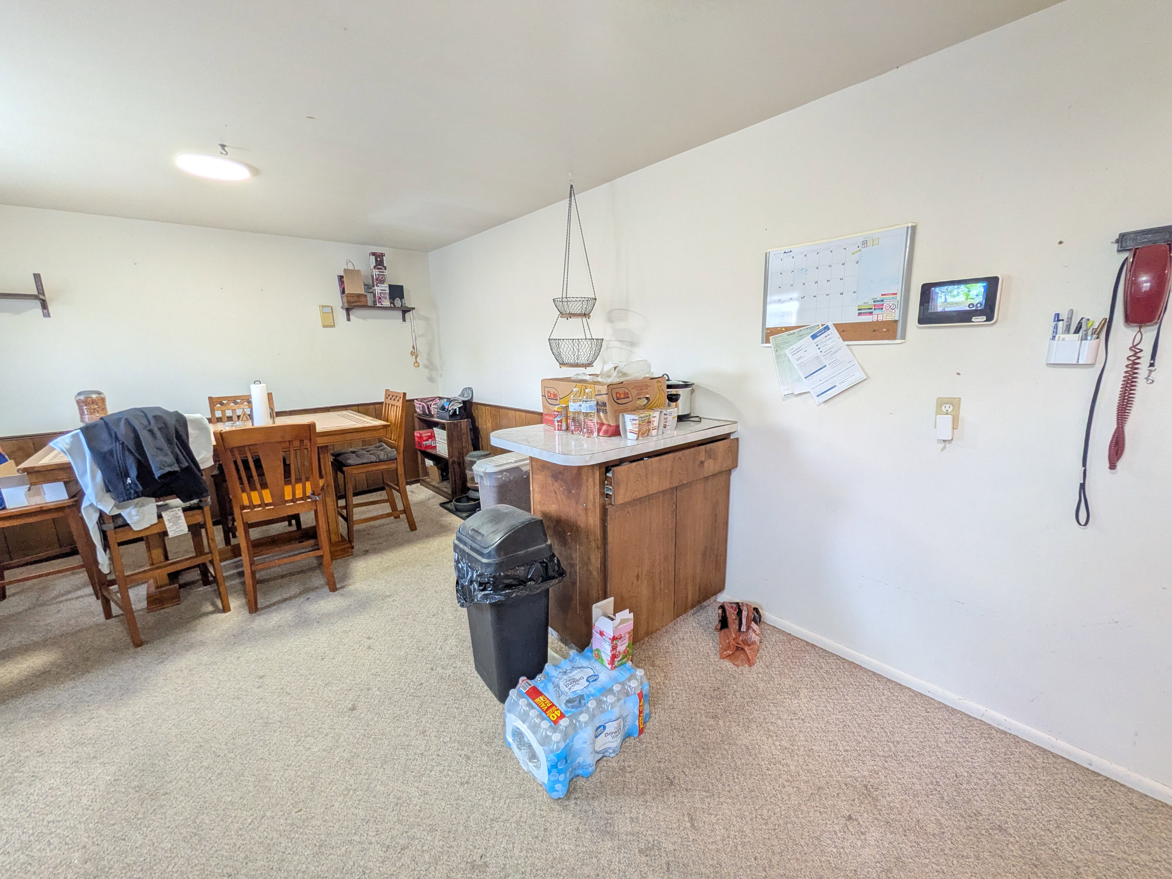 Carpeted room with a wooden dining table, a built-in wooden cabinet with a light-colored countertop, and a ceiling-mounted light fixture